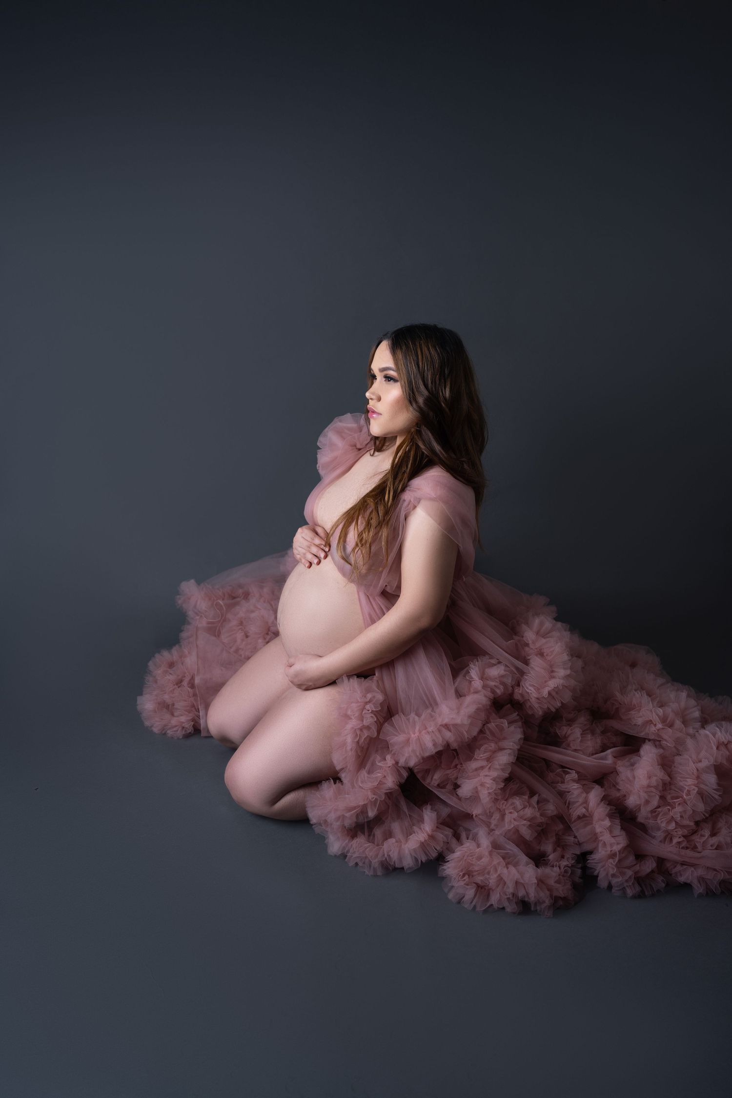 Pregnant woman in pink tulle robe kneeling on studio floor, cradling baby bump, looking sideways against gray background.