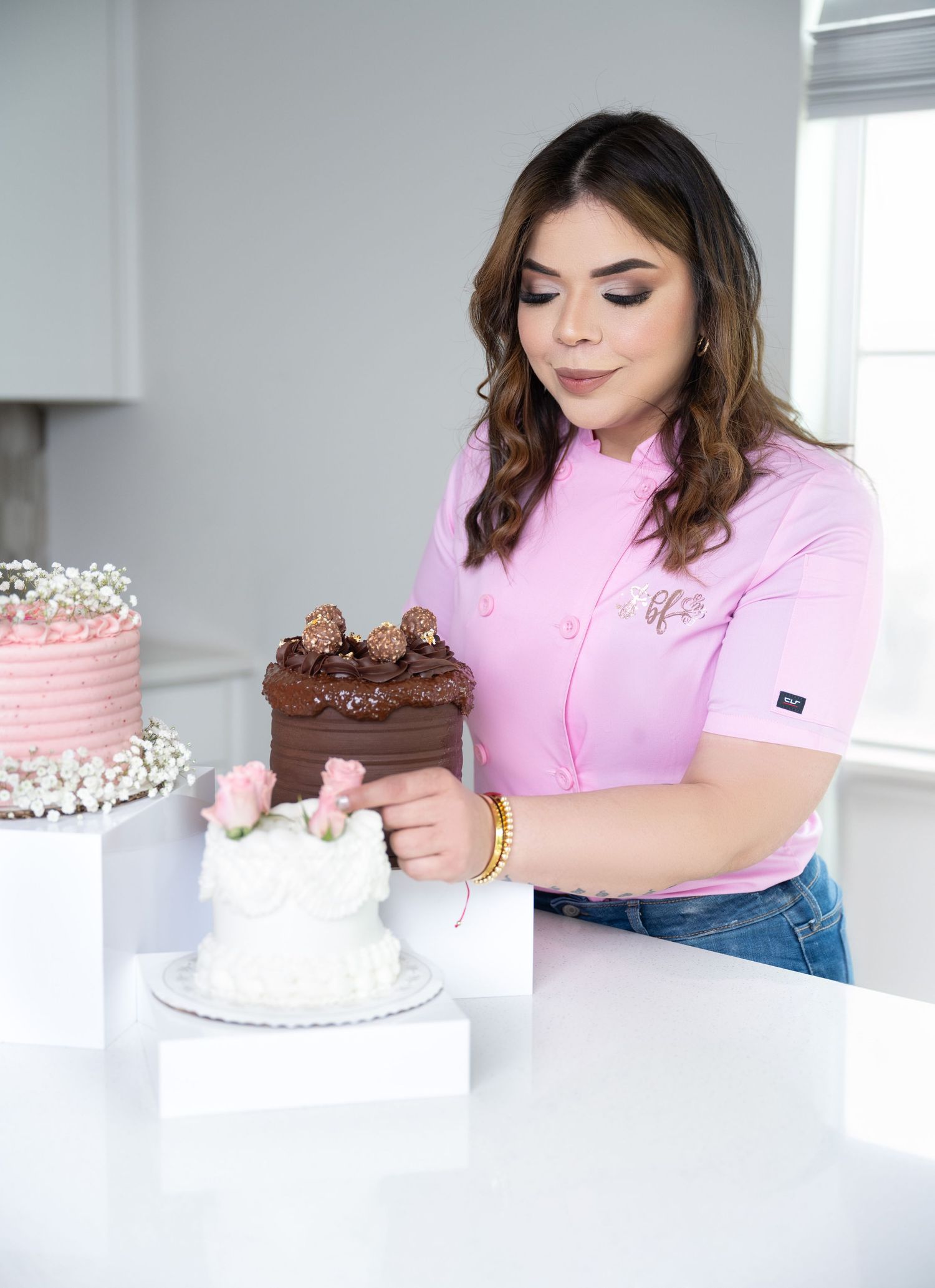 A female baker in a pink chef coat decorates chocolate and white cakes on a white counter in a bright kitchen.