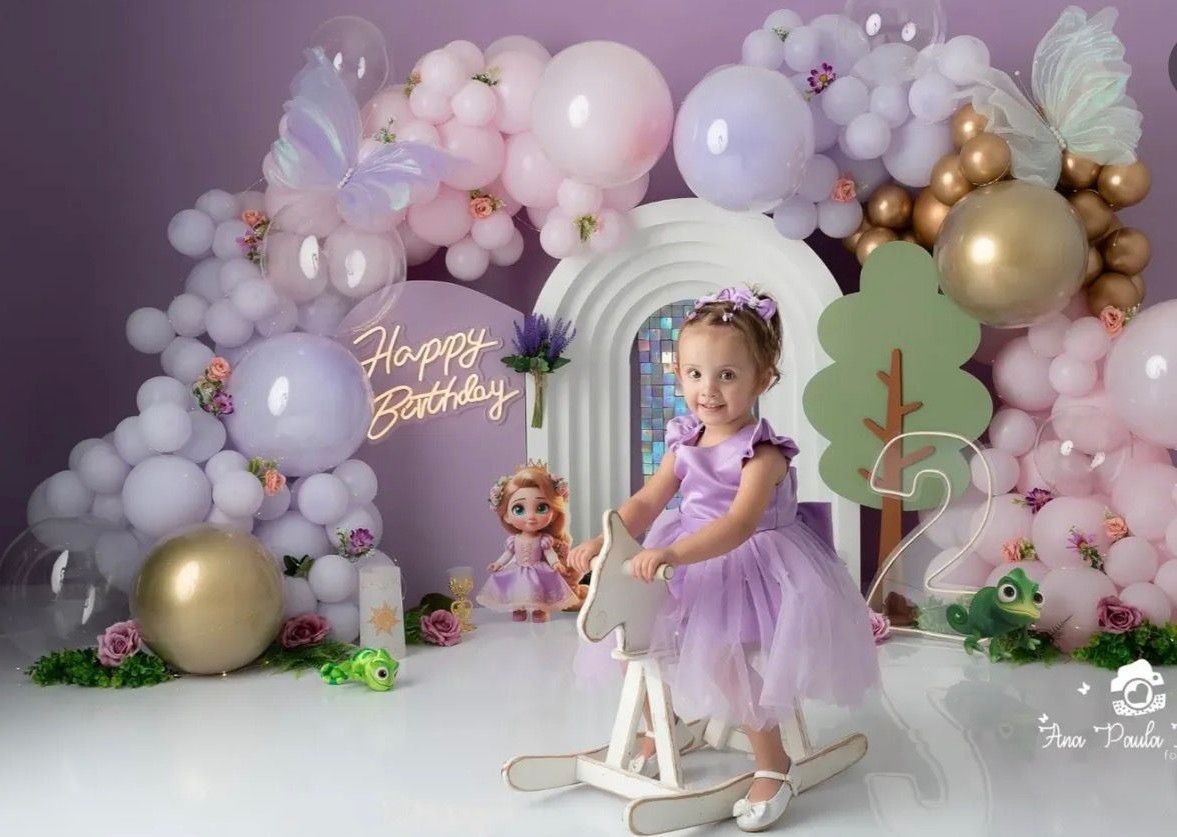 Toddler girl in purple tutu dress poses with rocking horse at birthday party with balloon arch backdrop.
