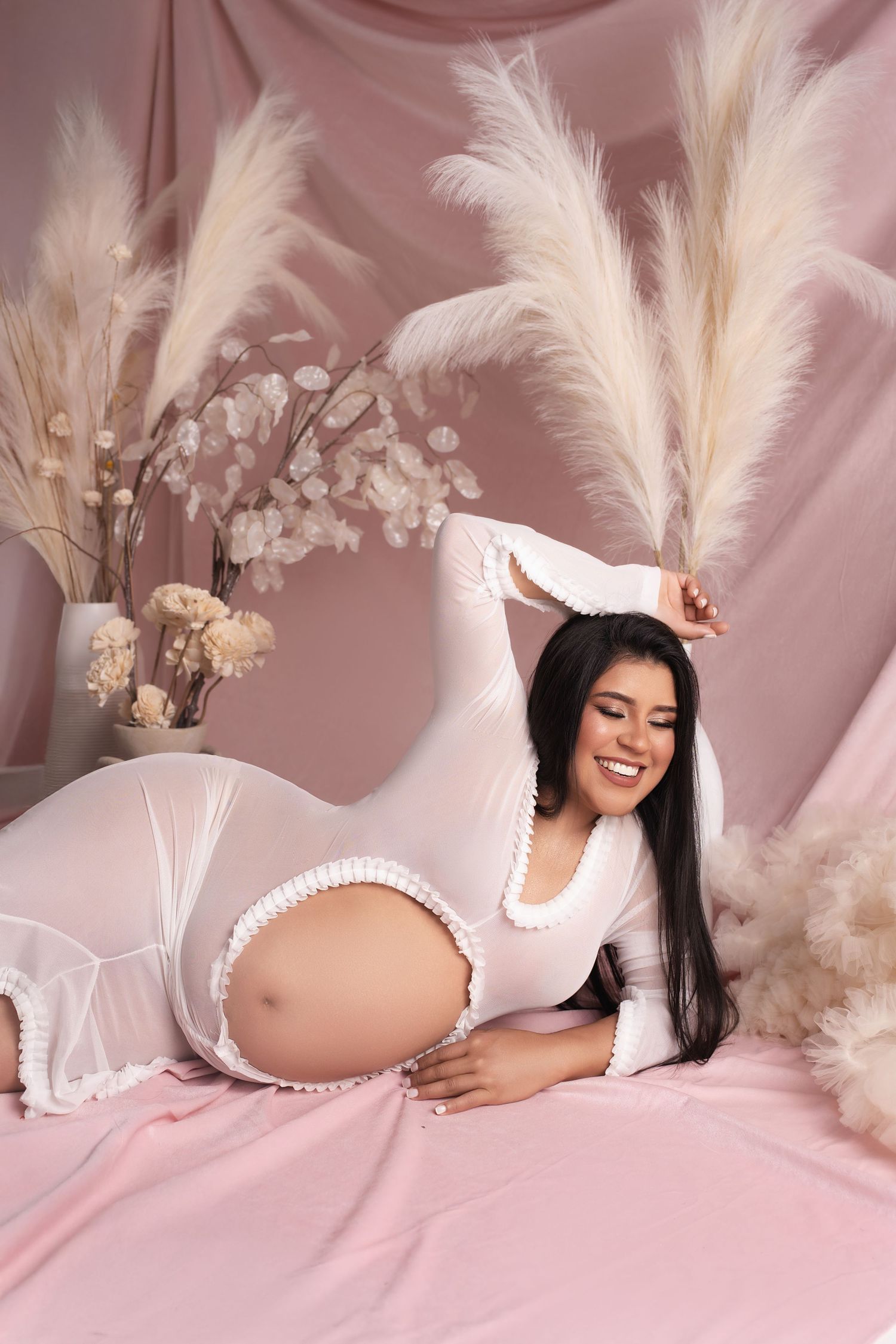 Maternity photoshoot featuring soft pink backdrop with pampas grass and floral decorations in romantic style.