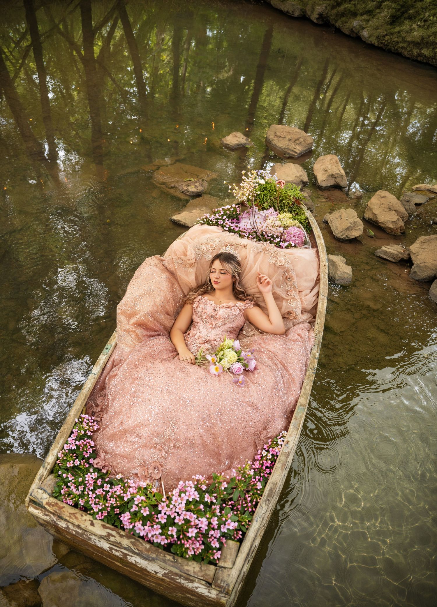 Young woman in pink ball gown lying in flower-filled wooden boat on serene forest river.