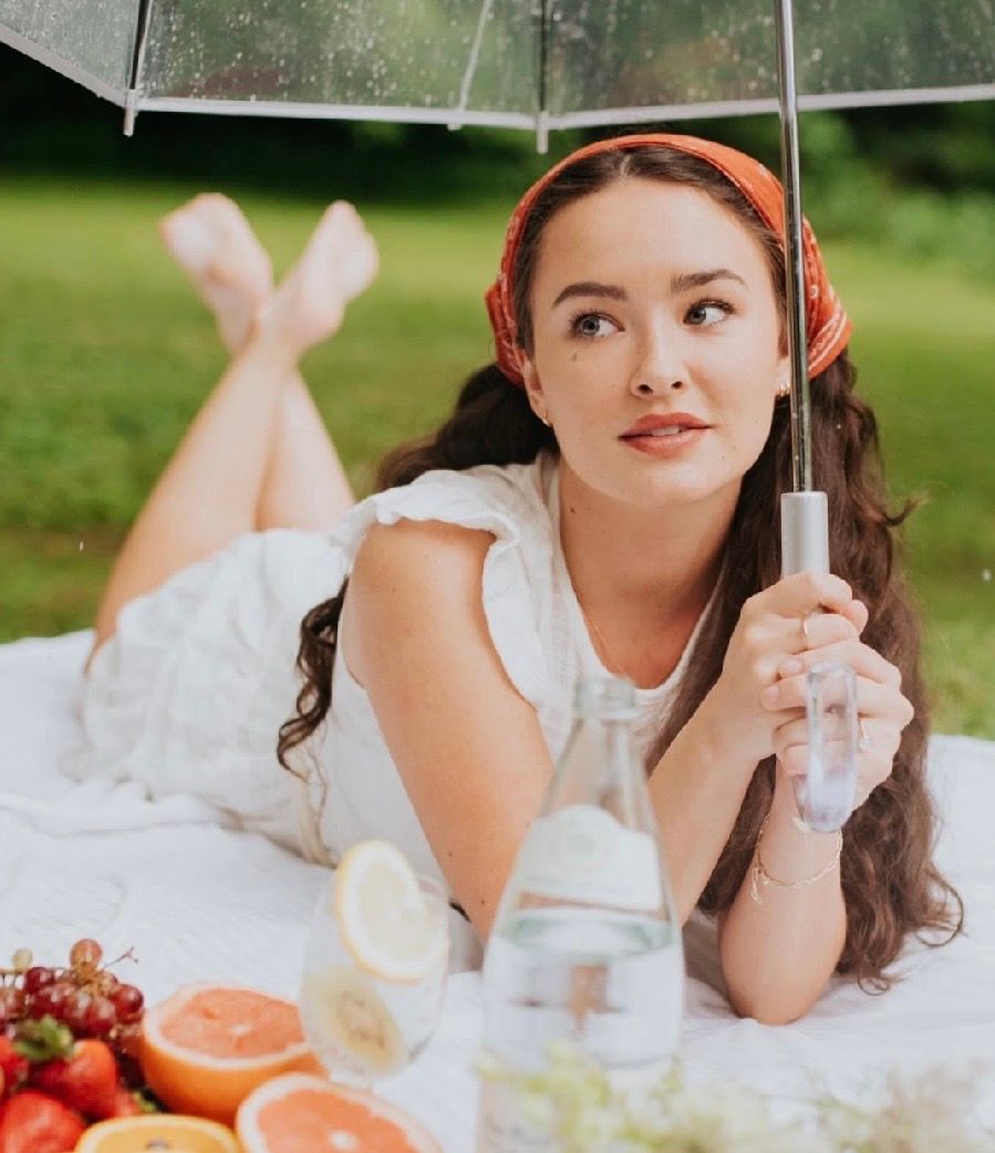 Young woman in white dress lying on blanket at picnic with fruits, holding clear umbrella, wearing red headband.