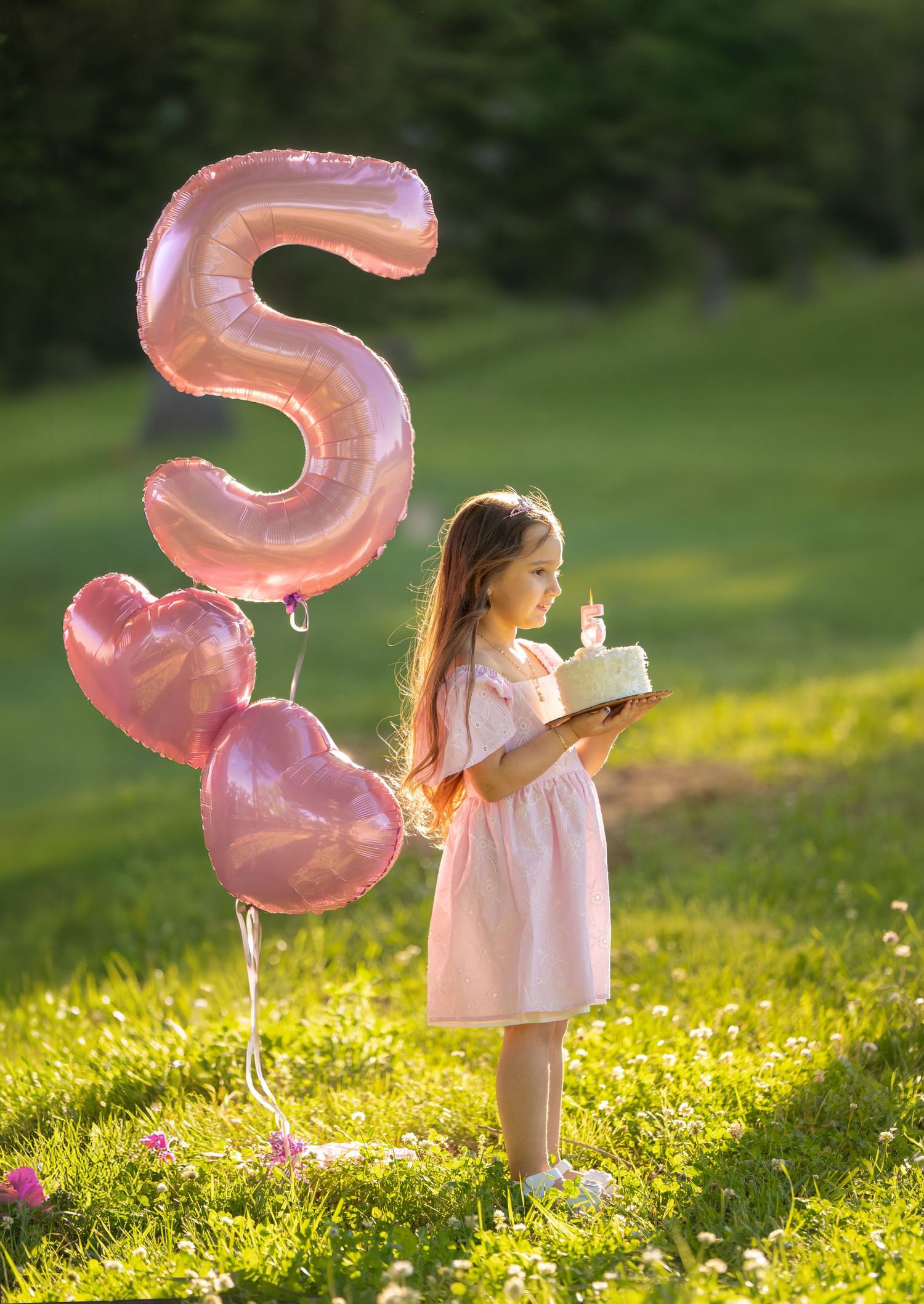 A little girl in a pink dress holds a birthday cake beside a number 5 balloon and pink heart balloons outdoors.