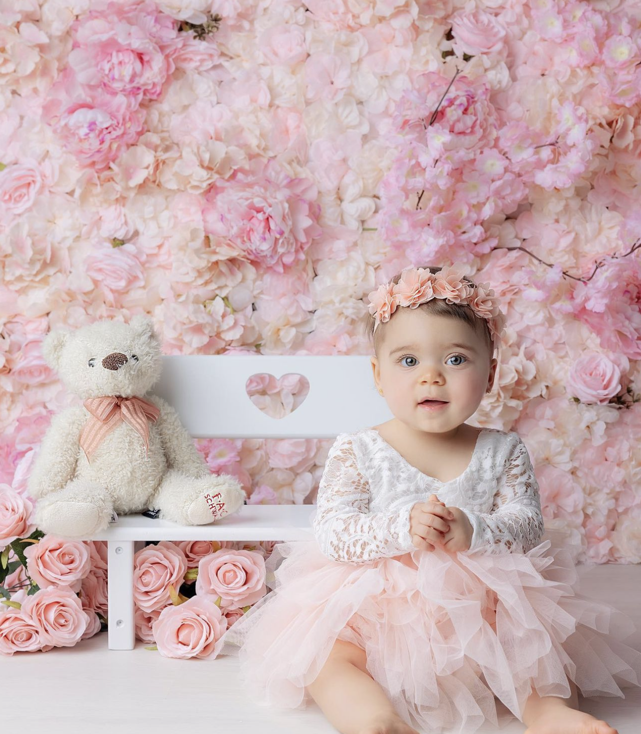 Baby girl in lace dress and floral crown sits on white bench beside teddy bear against pink floral backdrop.