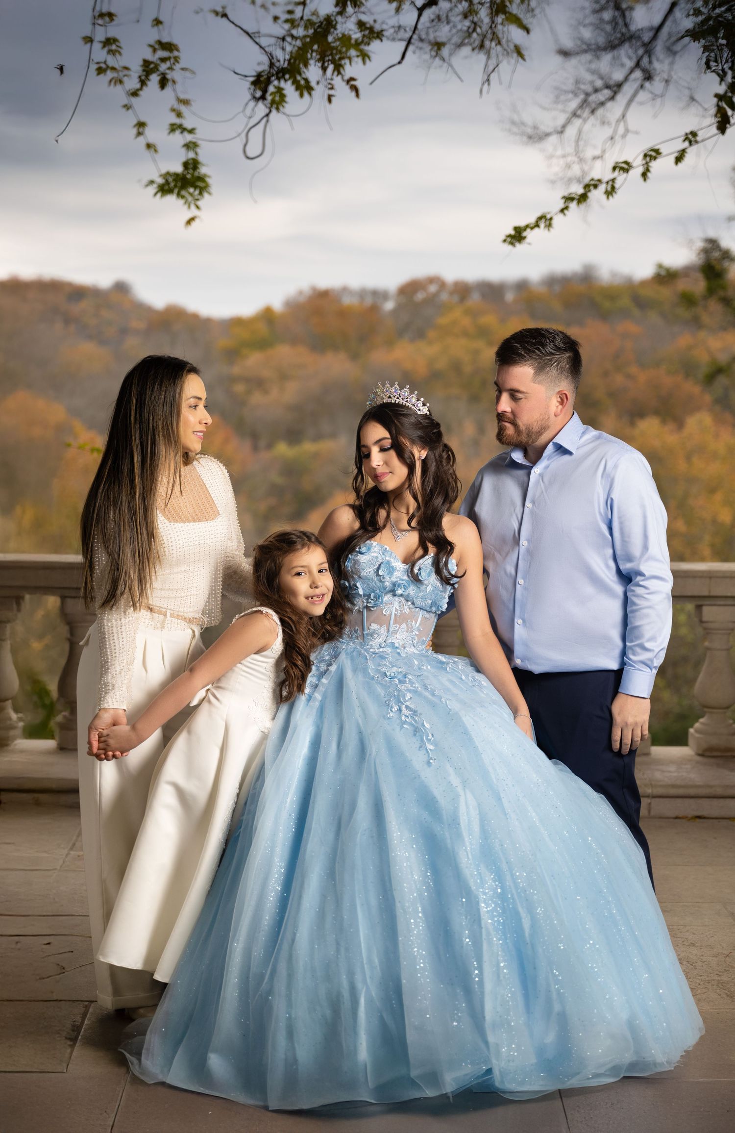 Family posing with quinceañera girl in blue ball gown and tiara on outdoor terrace with autumn trees.