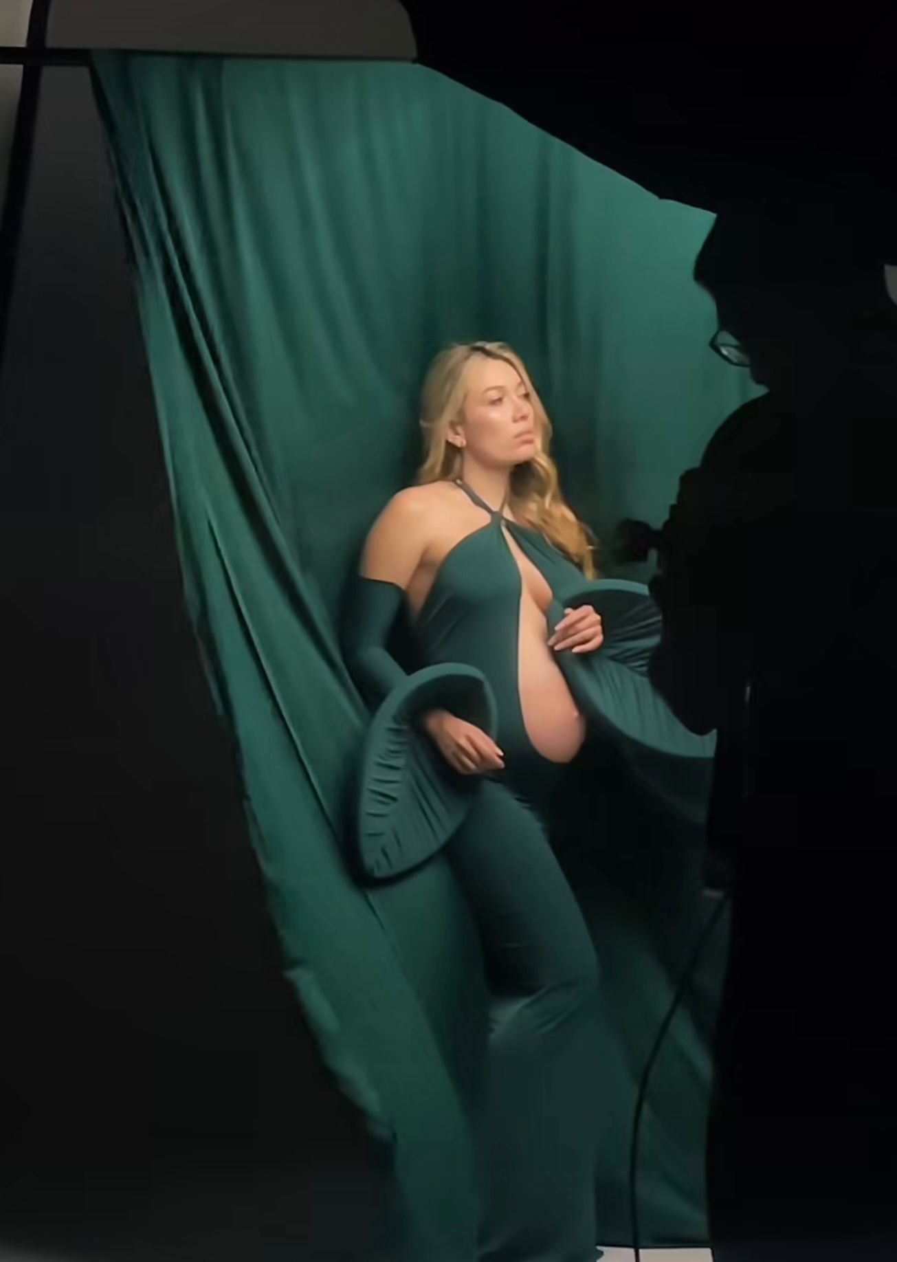 Pregnant woman in teal green maternity gown poses against matching fabric backdrop during professional photo shoot.