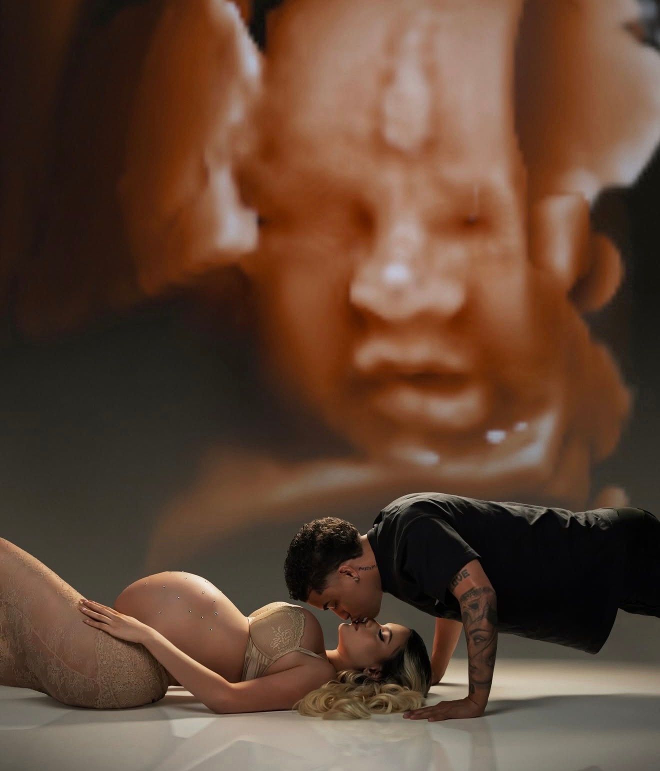 Two performers in an intimate theatrical scene lie on stage beneath a large projected face in dramatic lighting.
