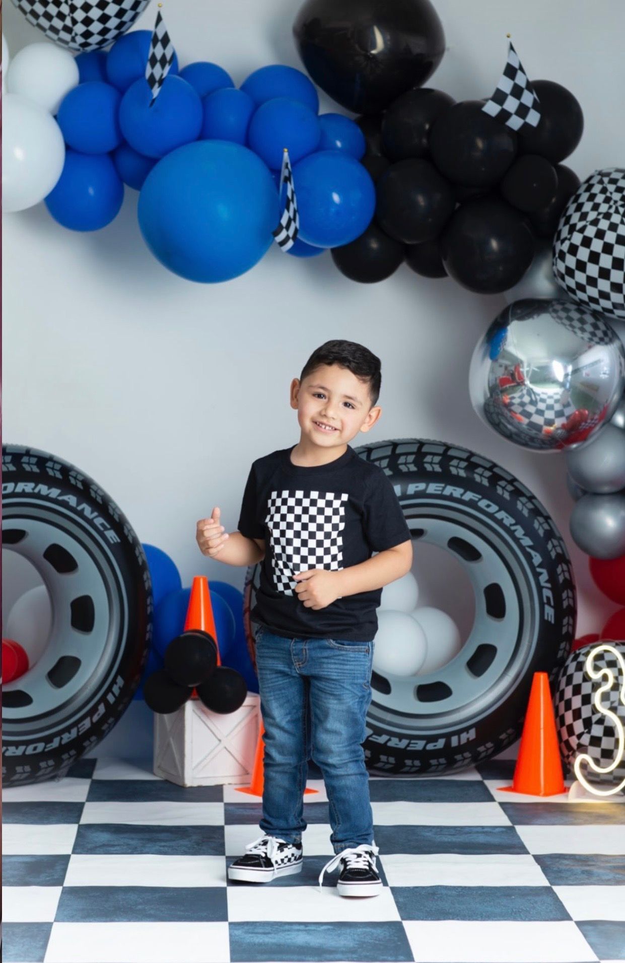 Smiling boy giving a thumbs up at a racing-themed birthday photoshoot with tire props, checkered floor, orange cones and balloons.