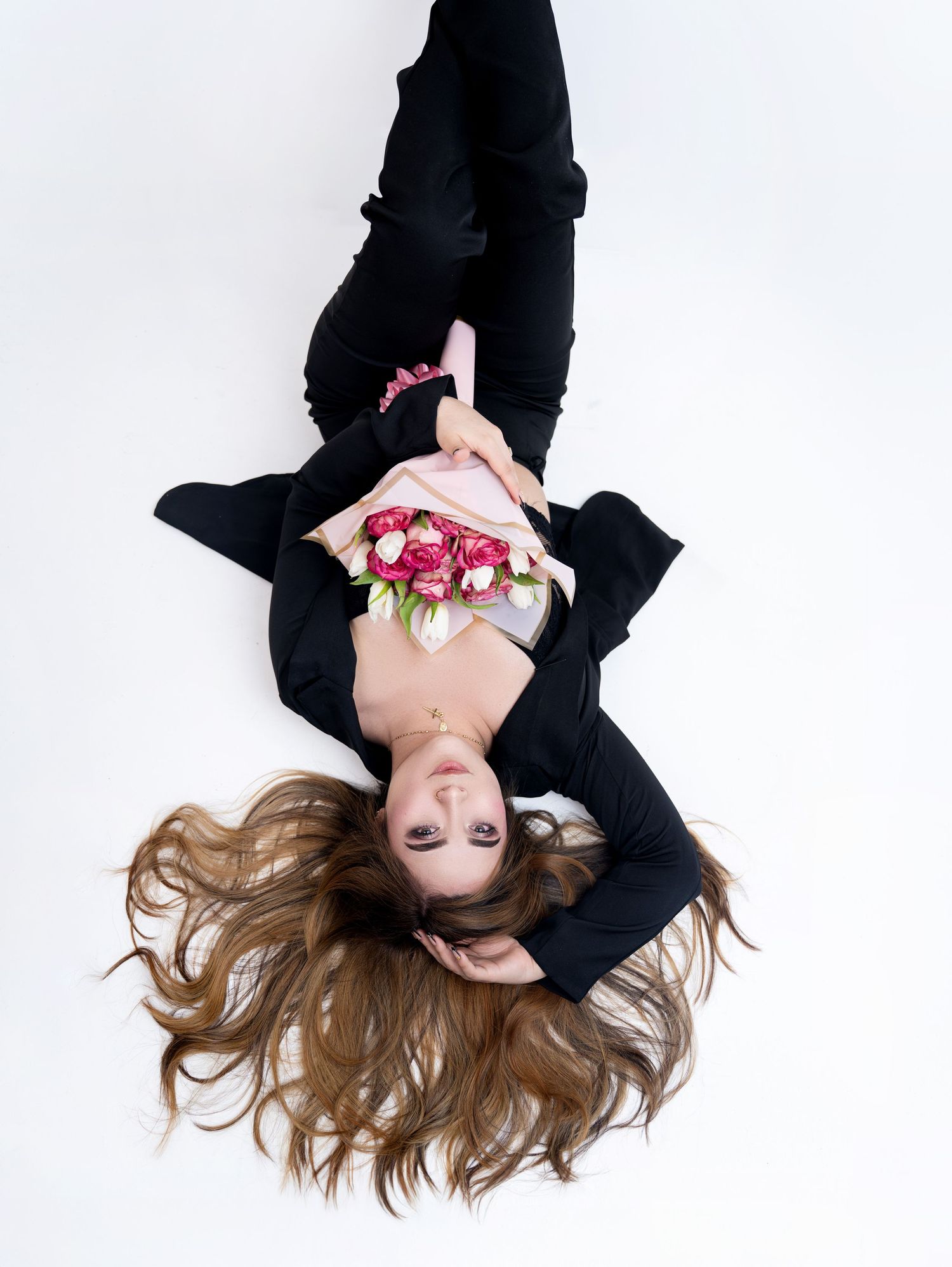 Woman in black outfit lying down holding pink and red flowers with long hair spread out on white background.