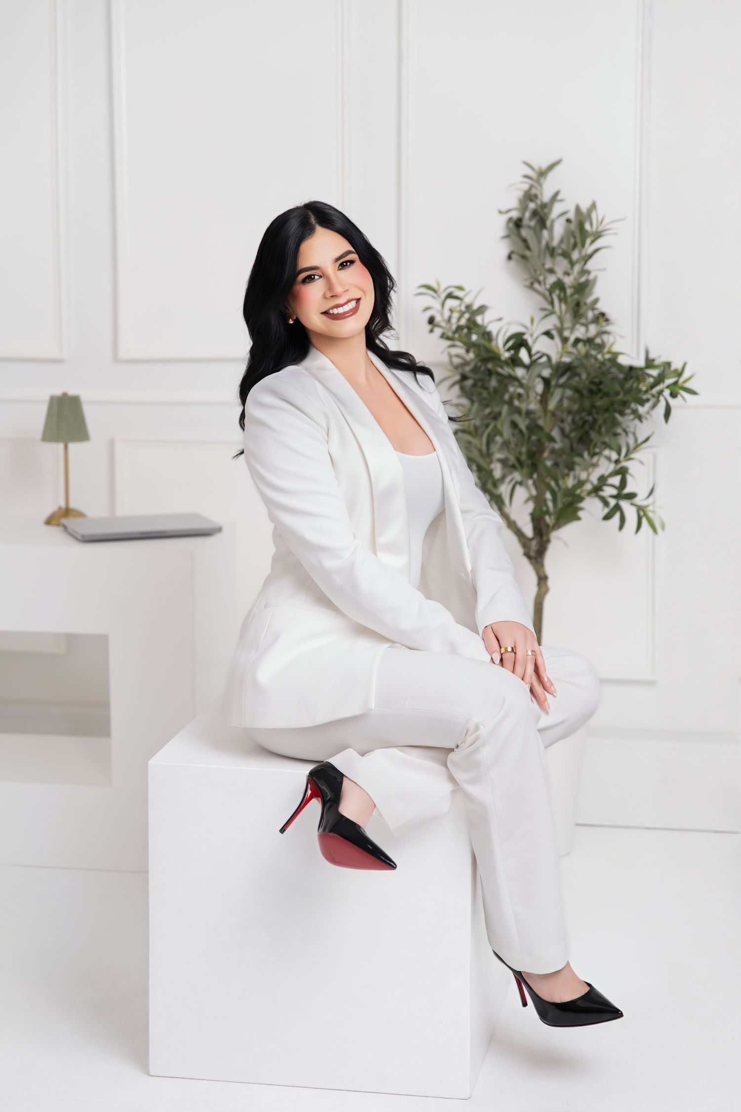 Woman in white suit and black heels sitting on white cube, smiling confidently in bright minimalist studio.