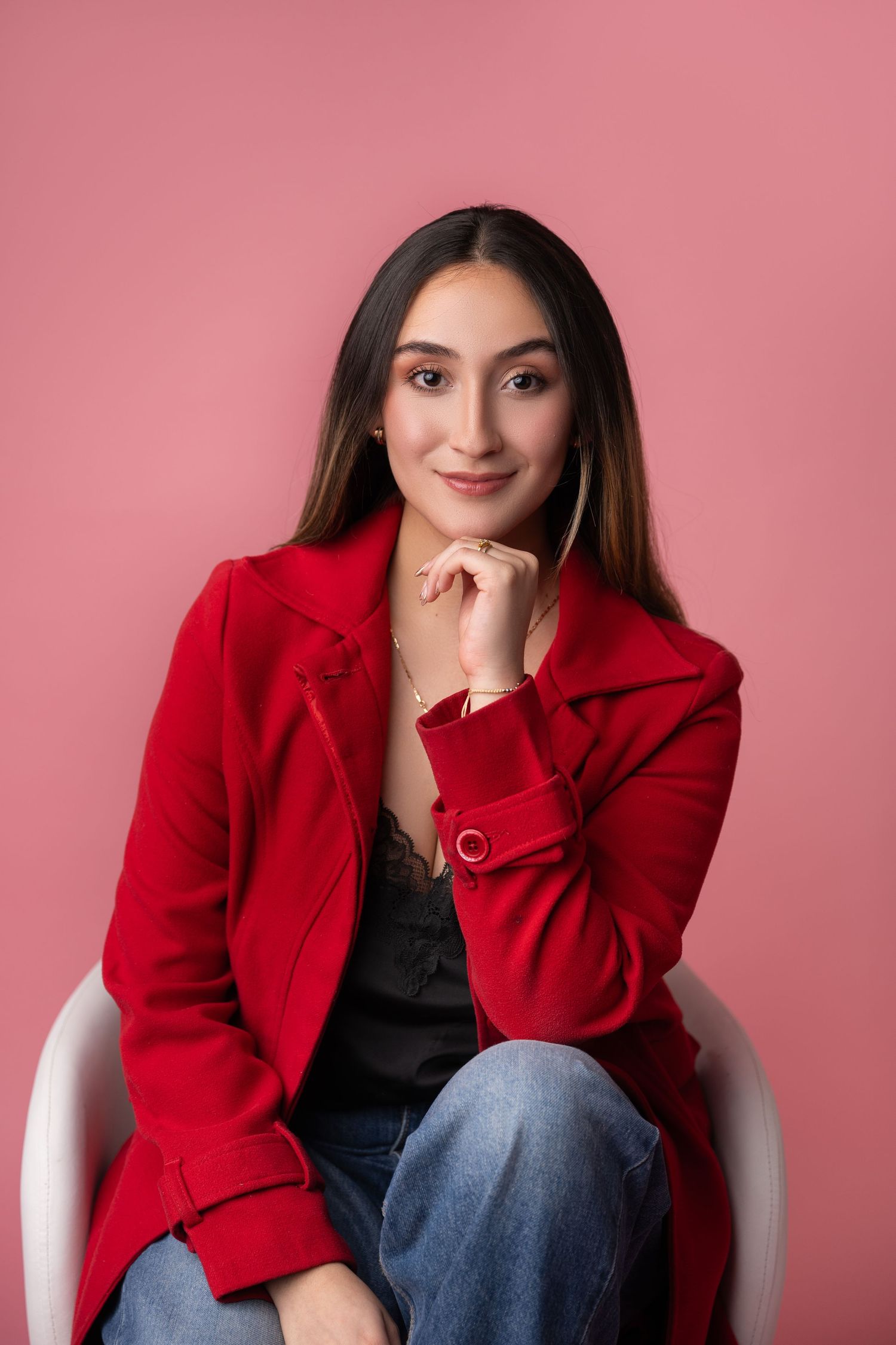 Young woman in red coat and jeans sits confidently on white chair against pink background, smiling at camera.