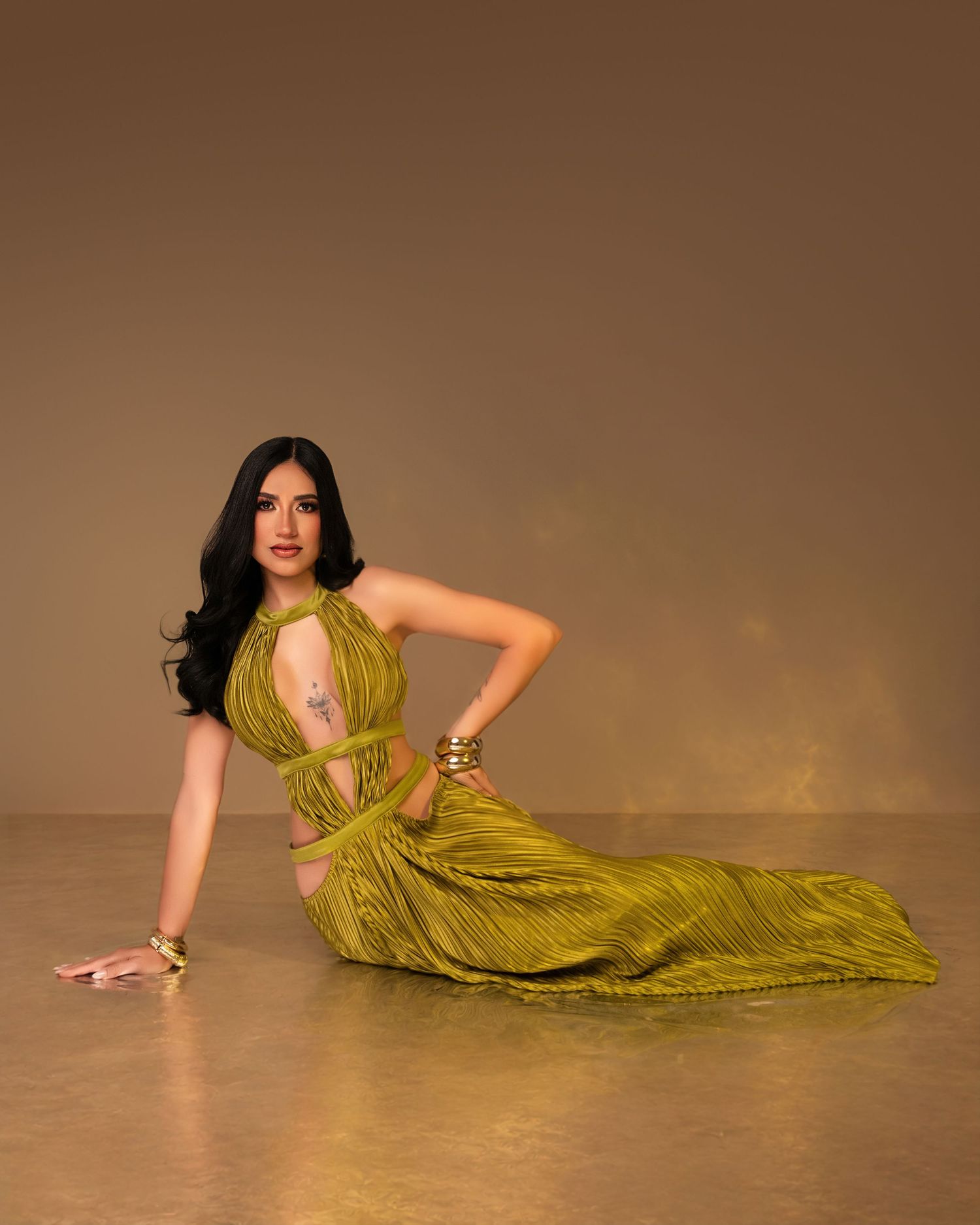 Woman in olive green halter dress poses gracefully on floor, wearing gold accessories against warm brown background.