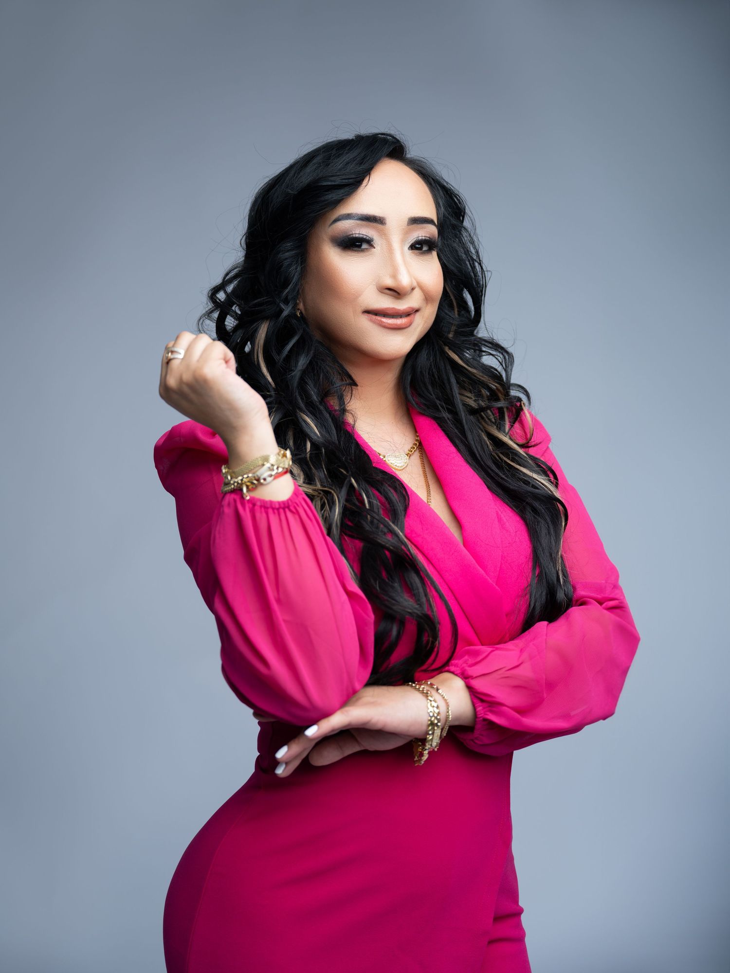 Woman with long wavy black hair posing confidently in a hot pink wrap dress with gold bracelets against a soft gray studio backdrop.