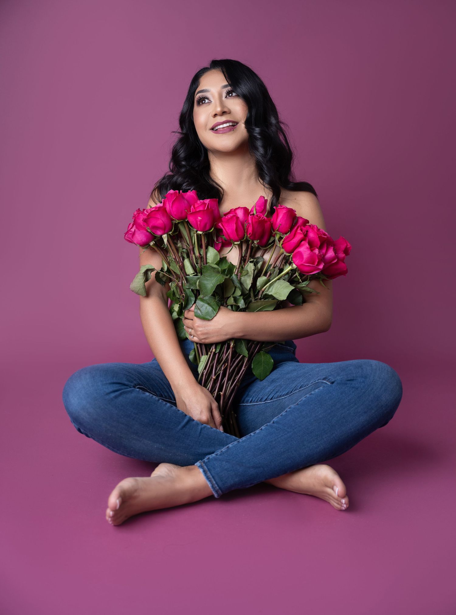 Smiling woman with long dark hair sits cross-legged in jeans, holding a bouquet of pink roses against a purple studio backdrop.