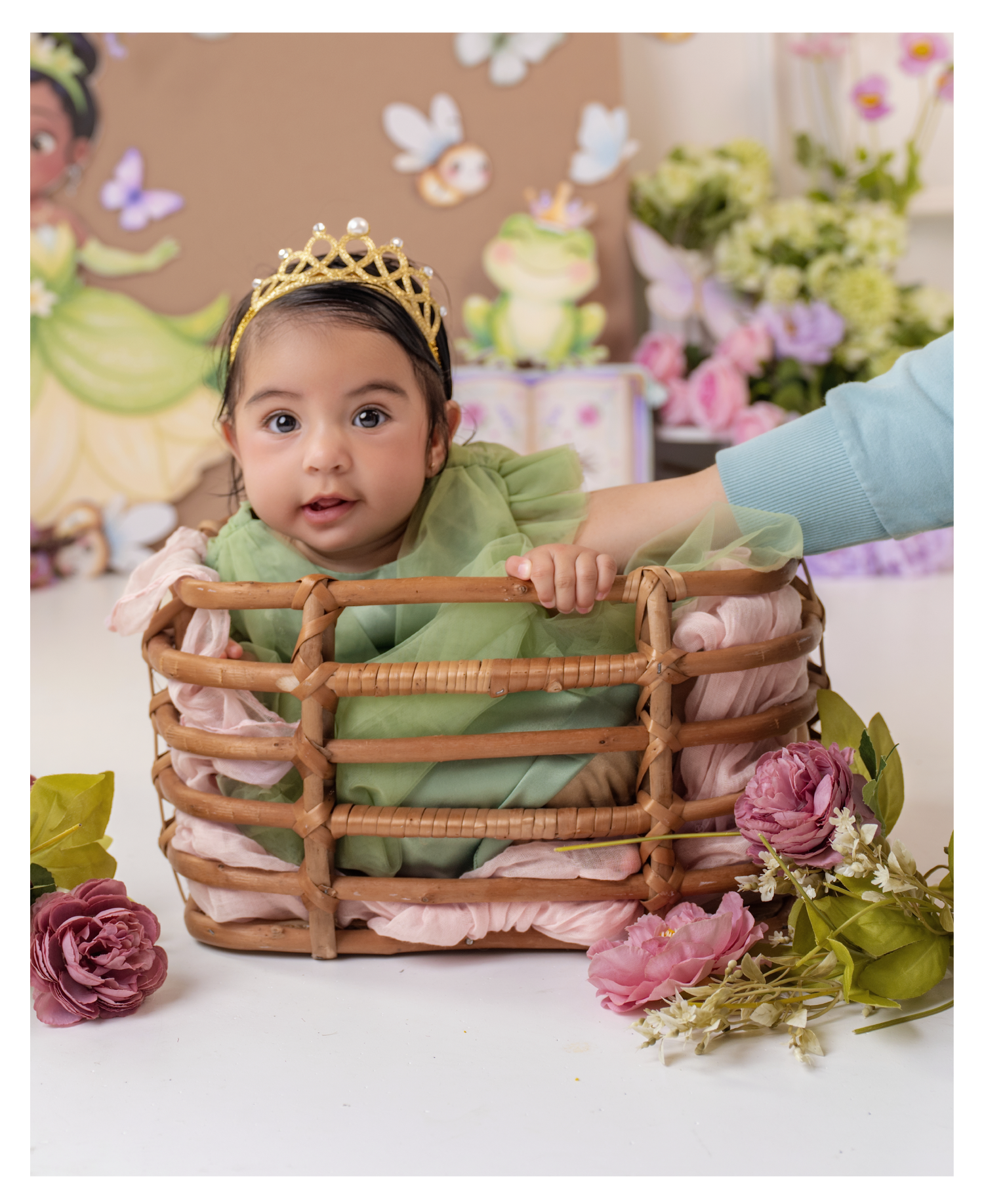 Baby girl in green dress and gold crown sits in a wicker basket surrounded by pink flowers.