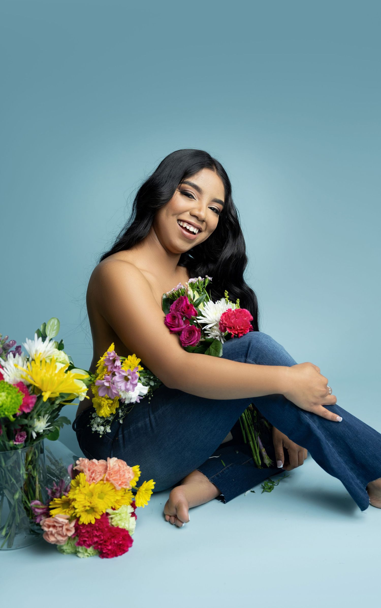 Smiling woman with long dark wavy hair sits barefoot in jeans holding fresh colorful flower bouquets against a soft blue studio backdrop.