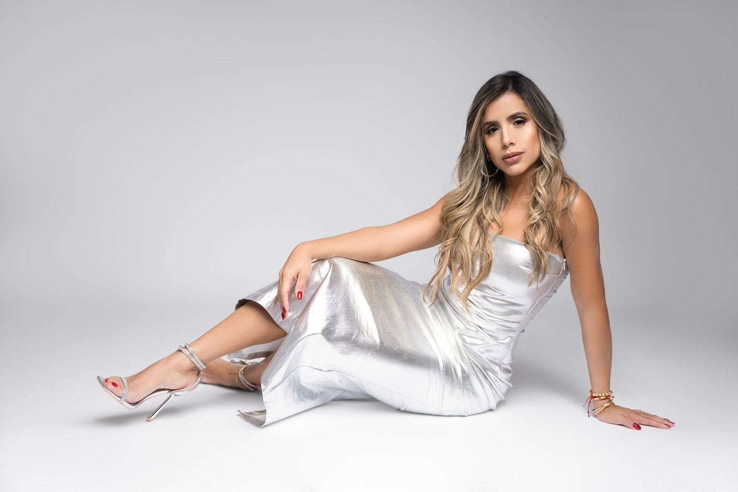 Woman with long wavy blonde hair poses seated on a white studio floor in a silver satin dress and strappy silver high heels.