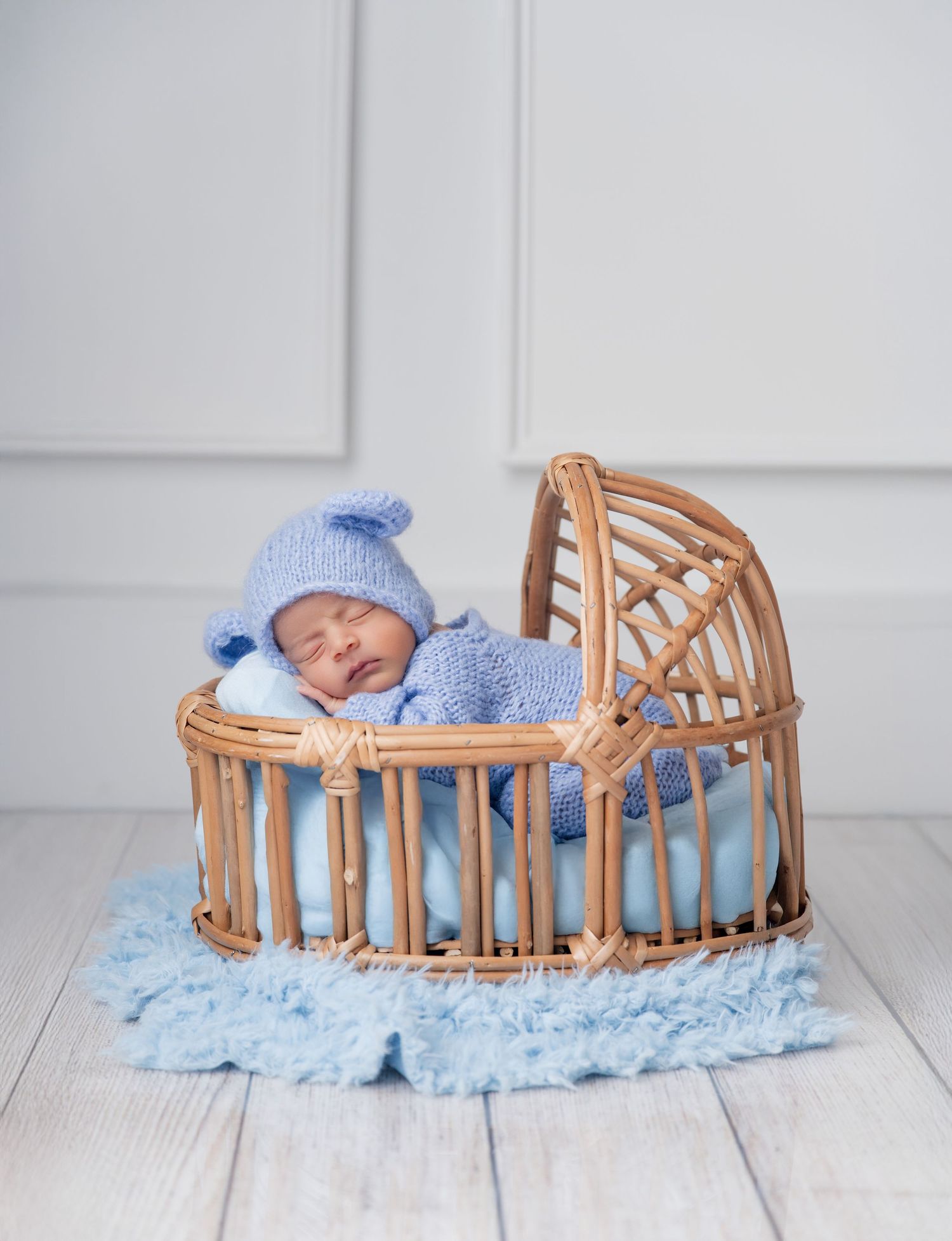 Newborn baby sleeping peacefully in a wicker bassinet, dressed in a blue knit outfit and hat.