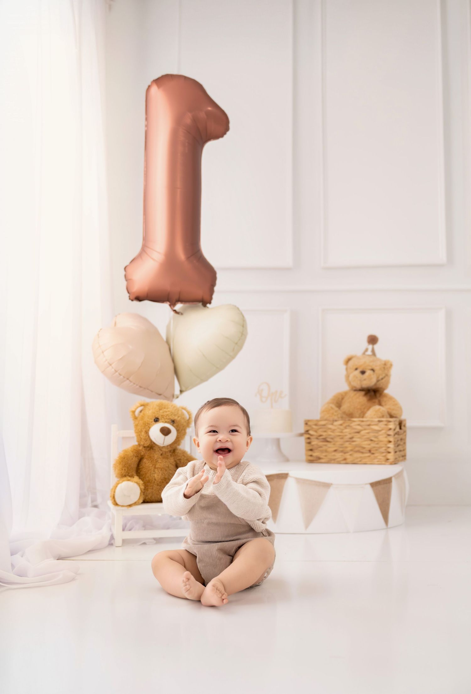 Happy baby sits on white floor celebrating first birthday with number 1 balloon and teddy bears in bright studio setting.