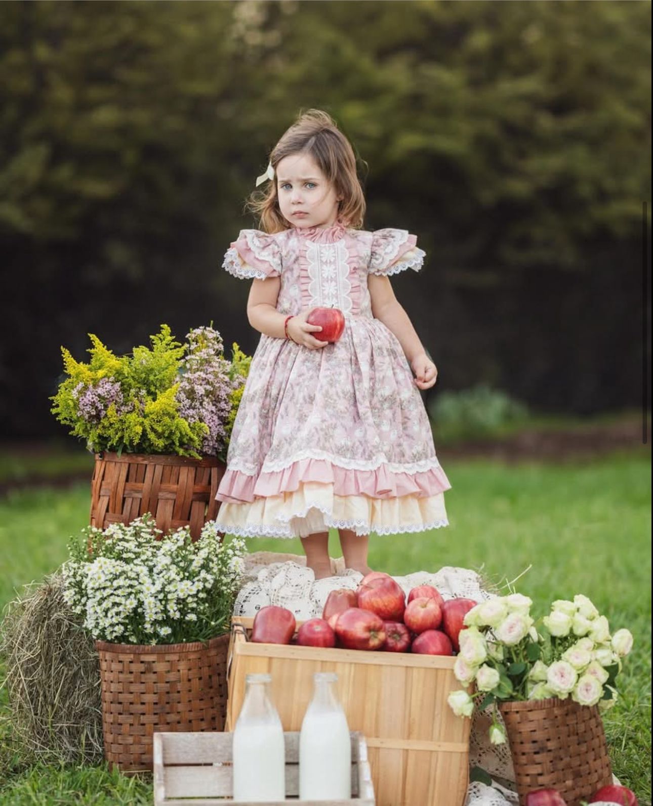 Young girl in floral dress holding apple, standing among baskets of apples, flowers, and milk bottles outdoors.
