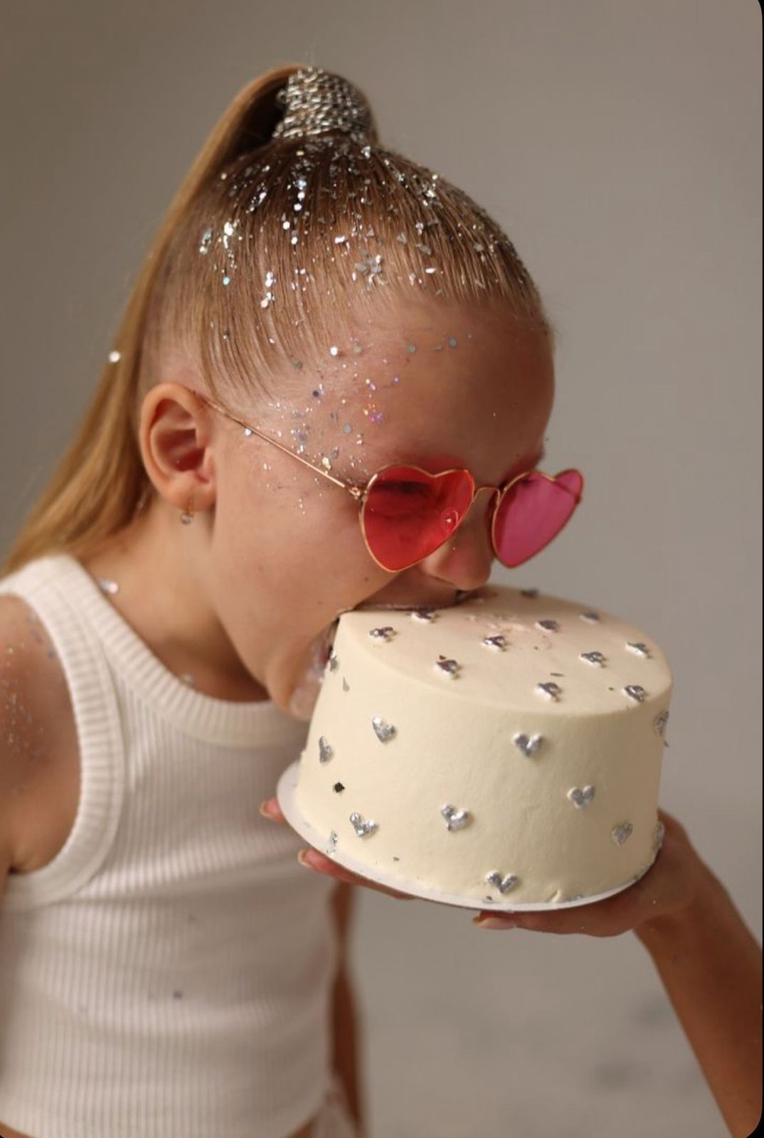 Young girl with glitter hair and pink heart sunglasses kissing a white cake decorated with silver hearts.