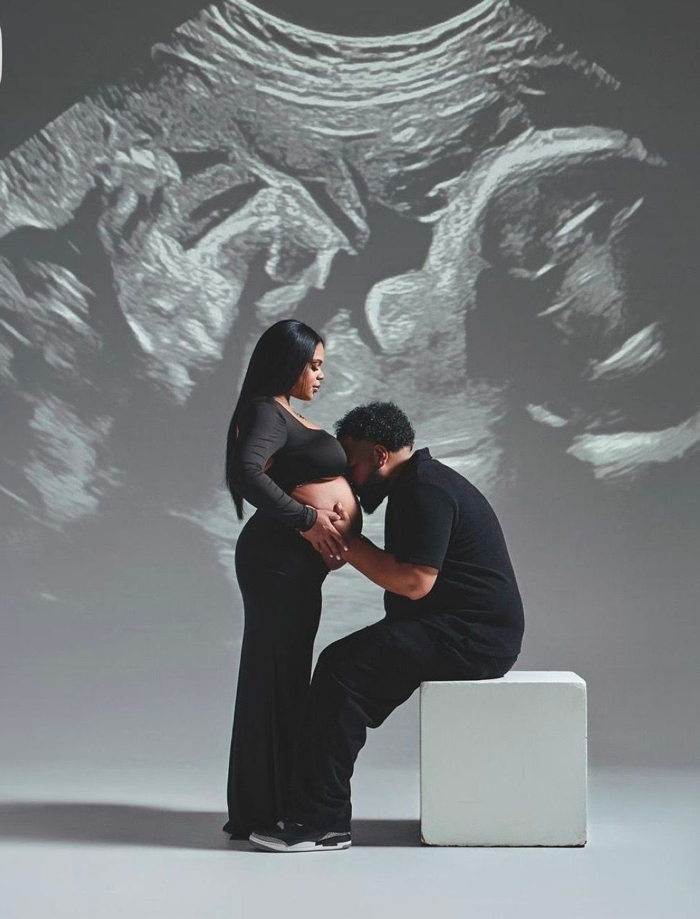 Couple during maternity shoot with ultrasound projection, man kissing pregnant belly in black outfits.