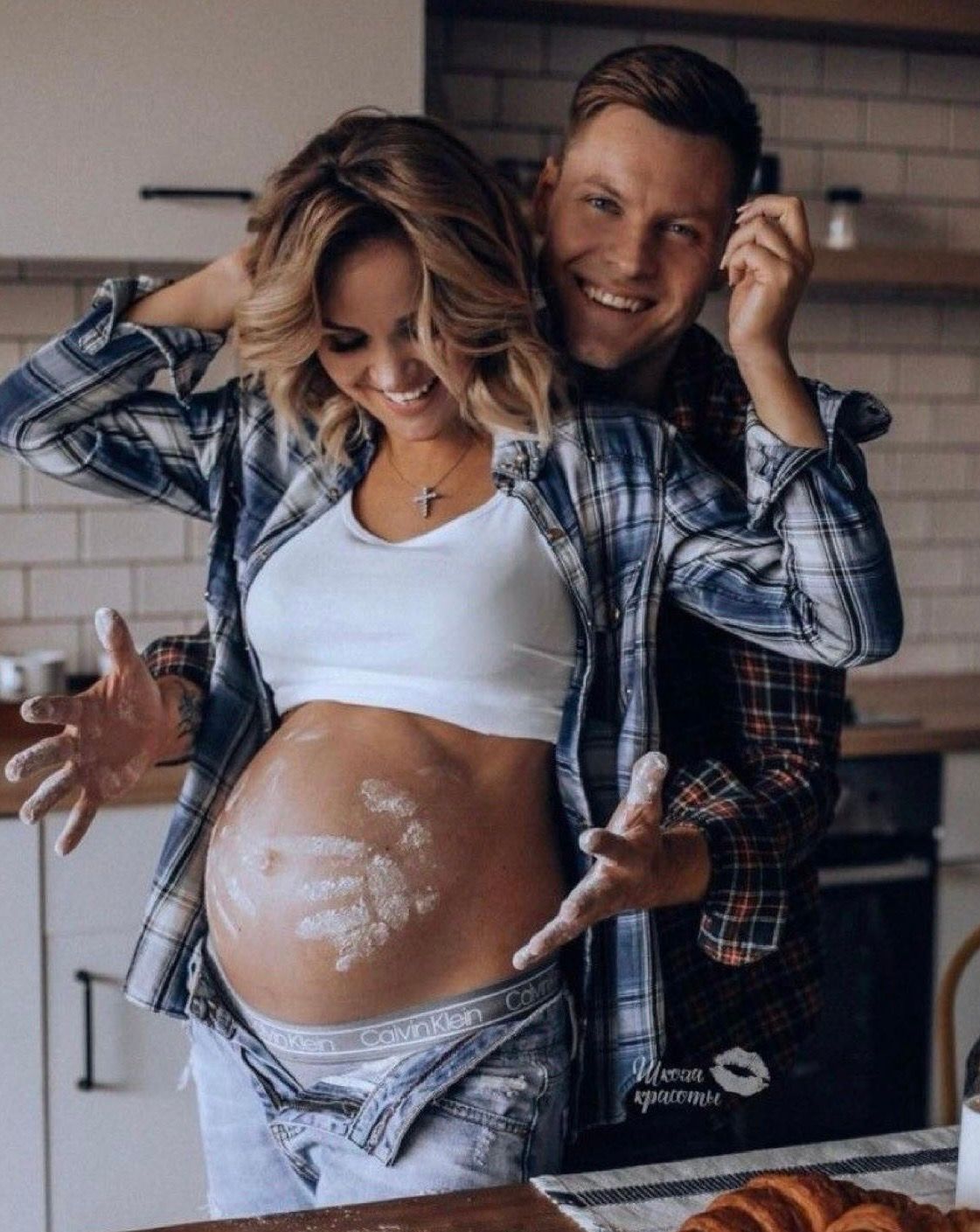 Happy couple in kitchen, pregnant woman with flour on belly, both smiling and wearing plaid shirts.