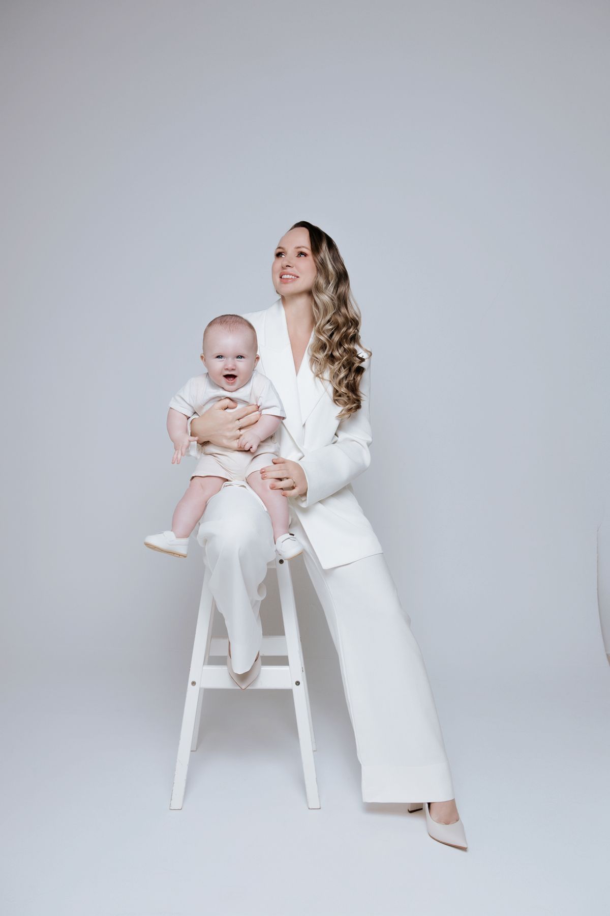 Woman in white suit sitting on stool holding smiling baby in pink outfit against white background.