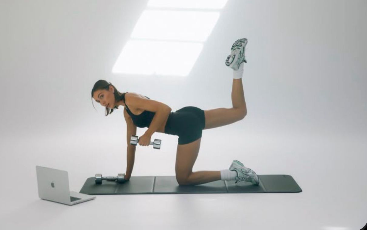 Woman performing donkey kick exercise on yoga mat with dumbbells, following laptop workout video in bright studio.