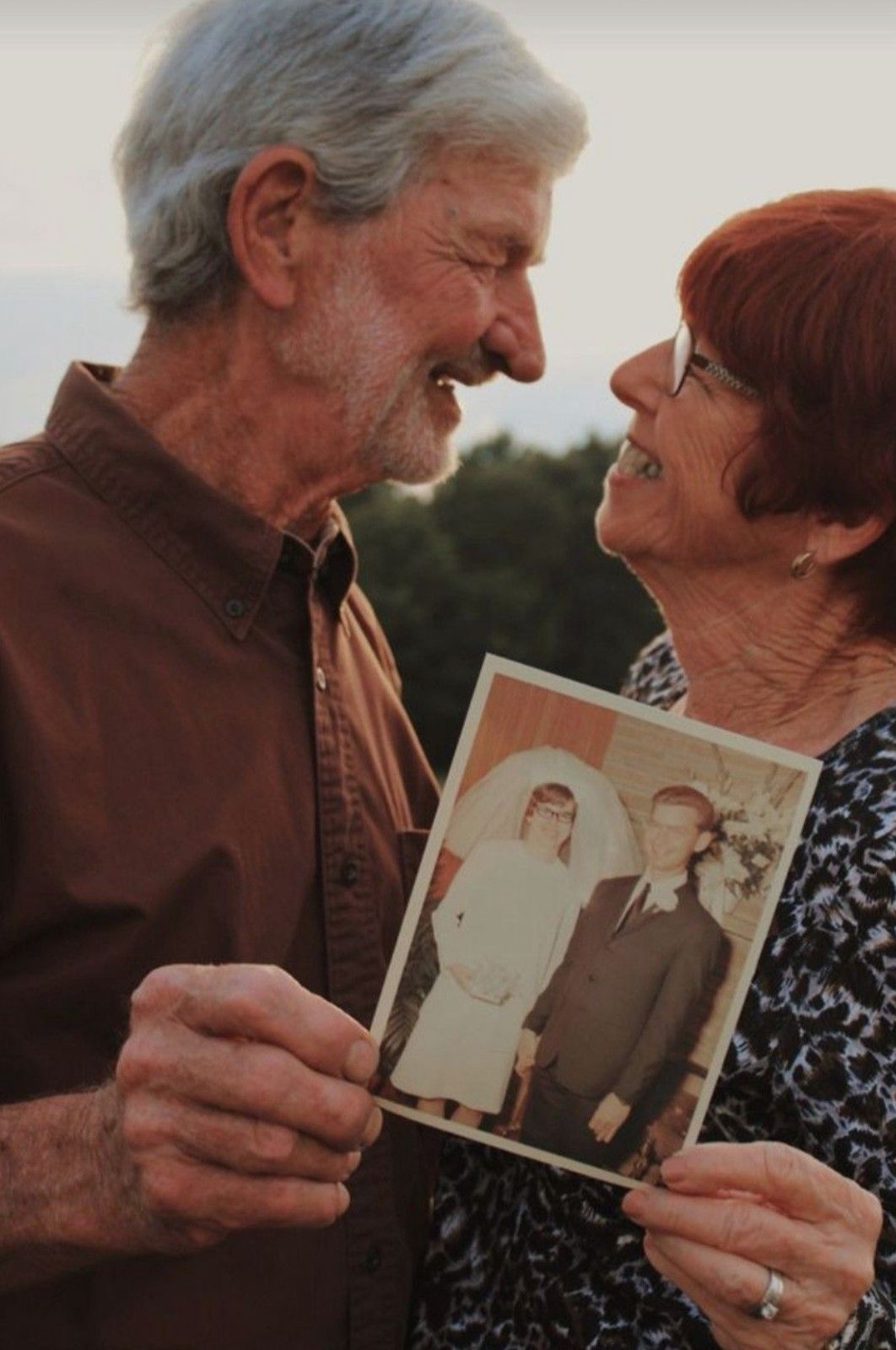 Elderly couple smiling at each other while holding an old wedding photo, celebrating their anniversary outdoors.