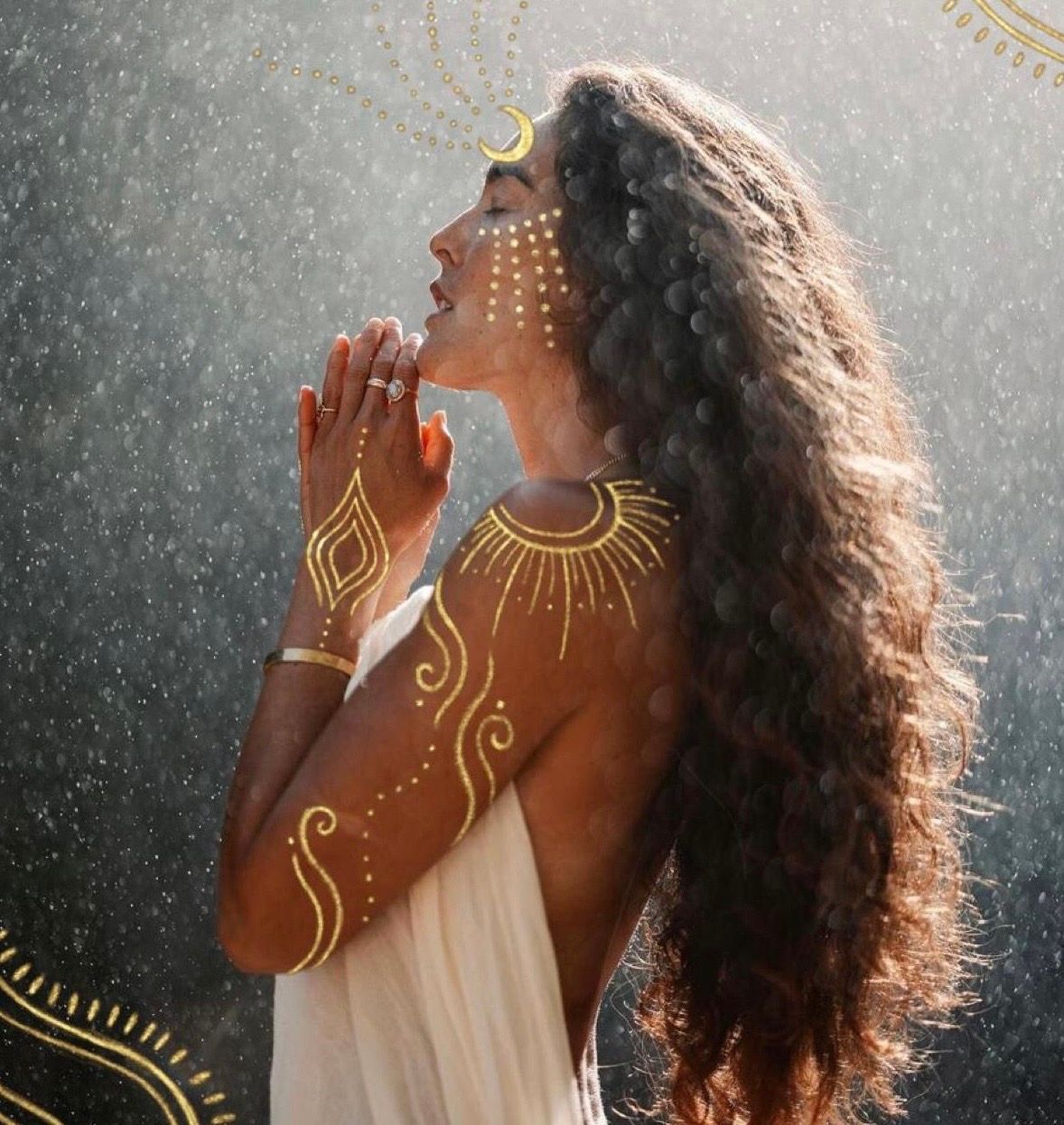 Woman with golden body paint and crescent moon headpiece praying in rain, wearing white dress.