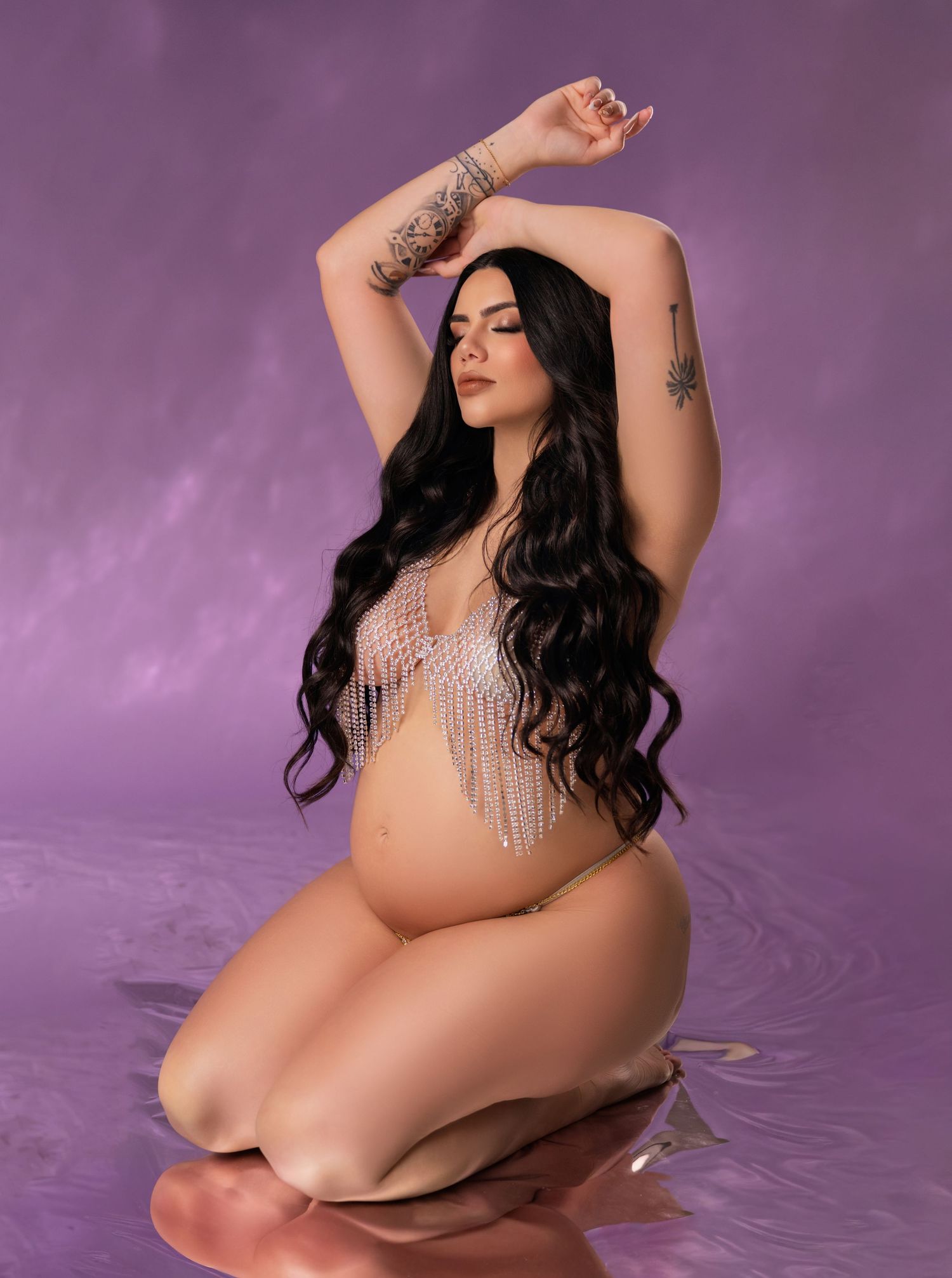 Pregnant woman with long dark hair poses gracefully in crystal bodypiece against purple backdrop.