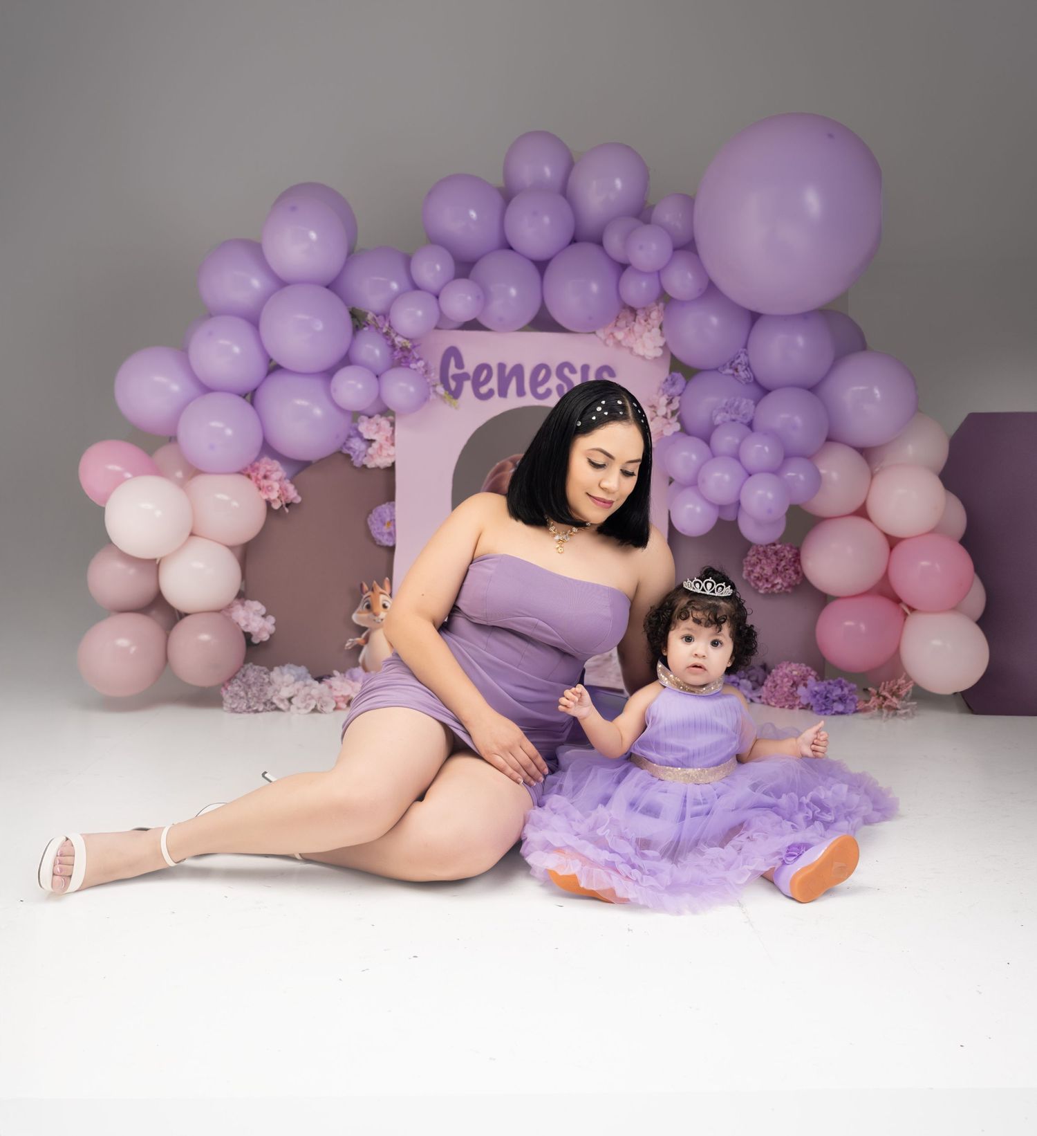 Mother and toddler in matching purple dresses pose before a lavender balloon arch with the name Genesis at a birthday celebration.