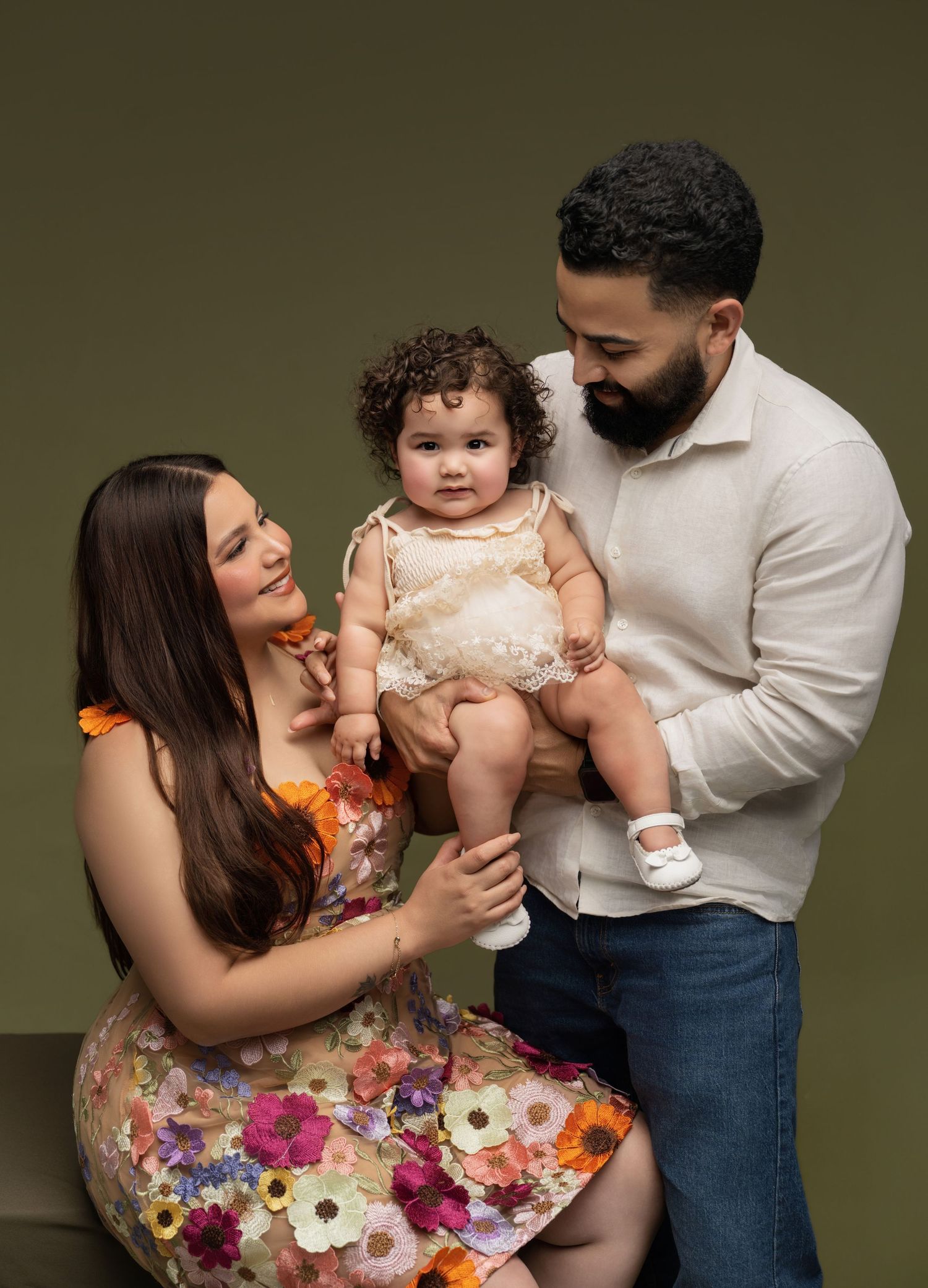 A happy family with a mother in a floral dress and father in white shirt holding their smiling baby girl in a studio.