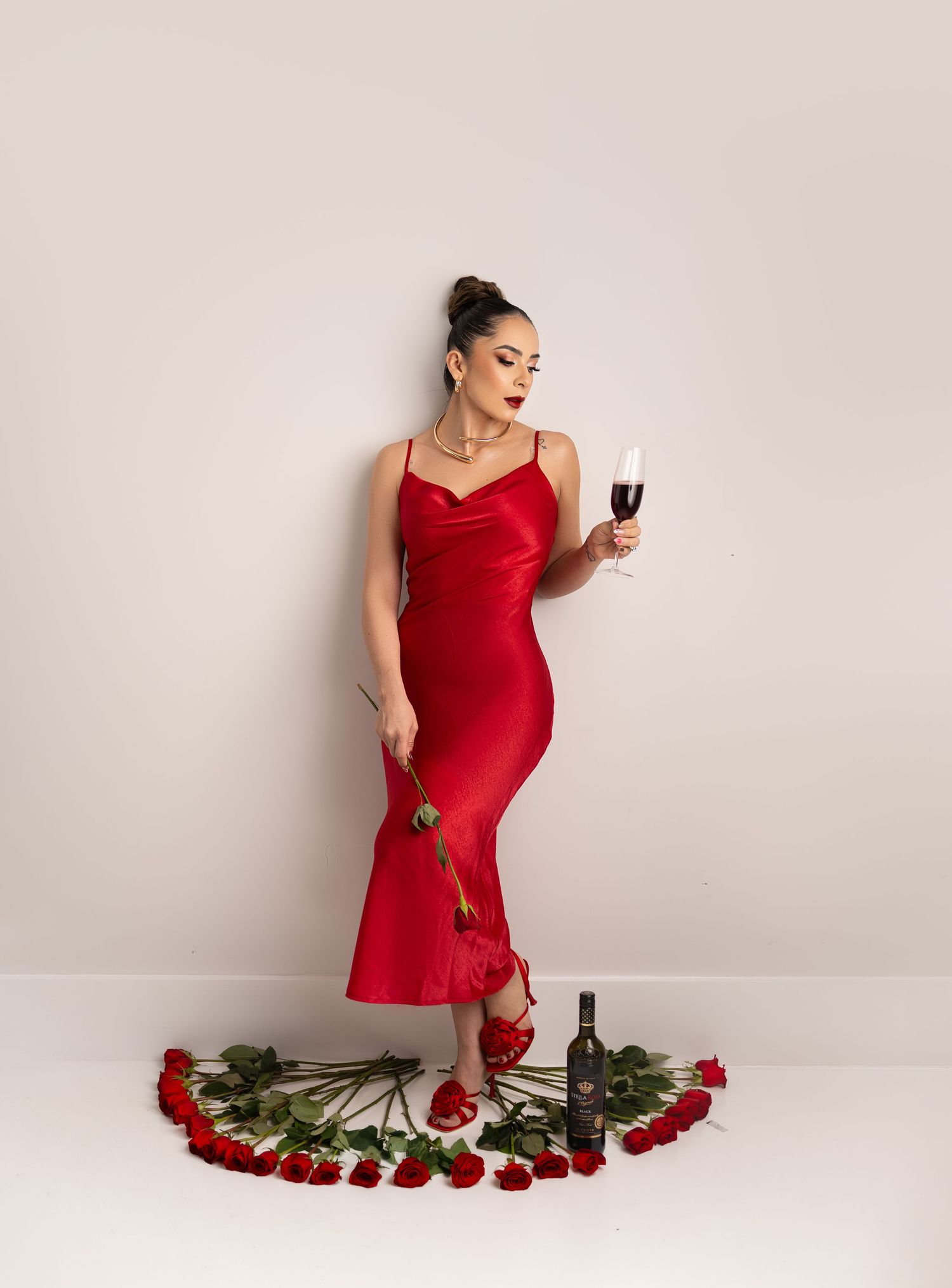 Person in a red satin dress poses elegantly with wine glass surrounded by scattered red roses on the floor.