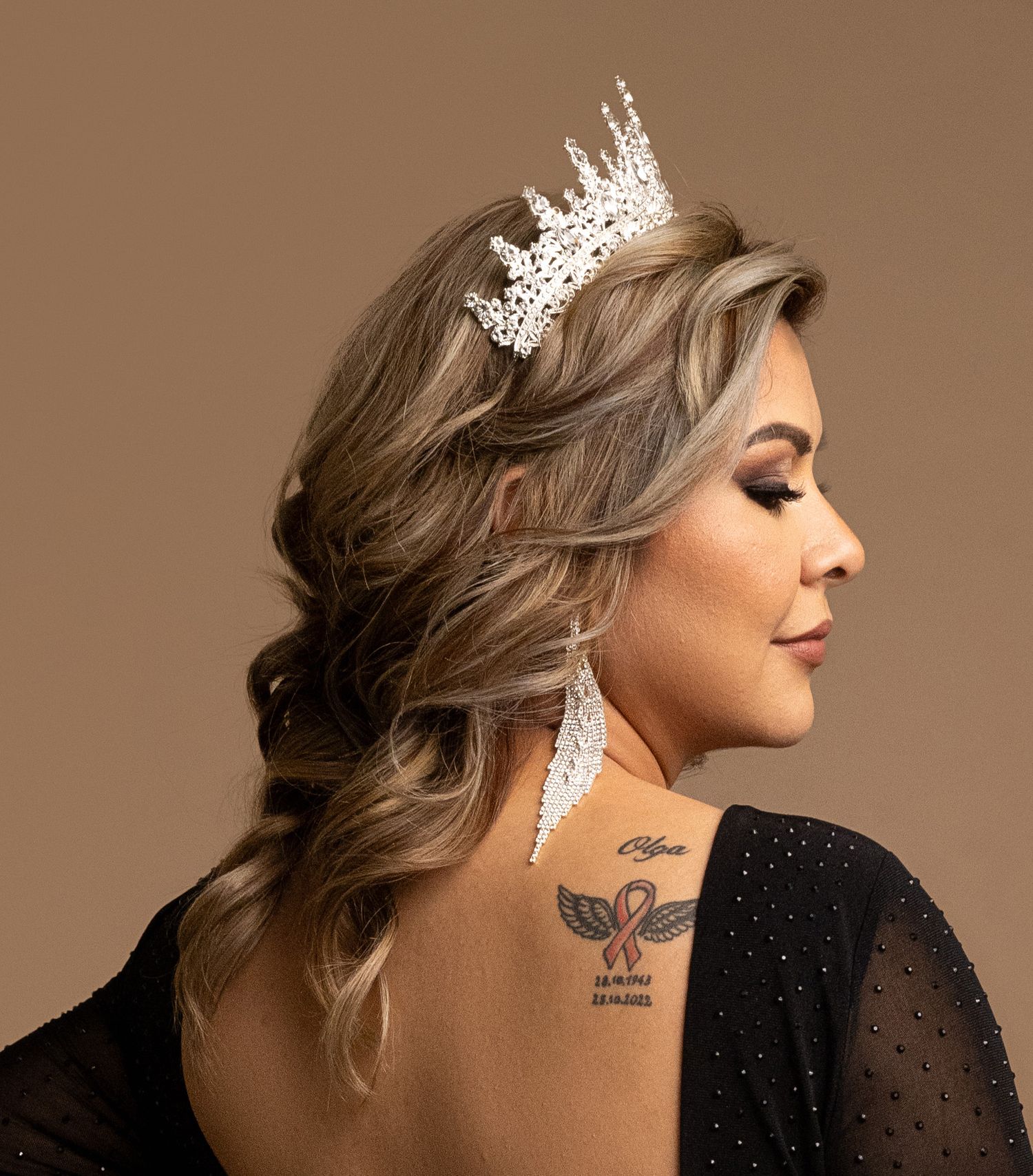 A glamorous portrait featuring a crystal crown and shoulder tattoo against a brown backdrop.