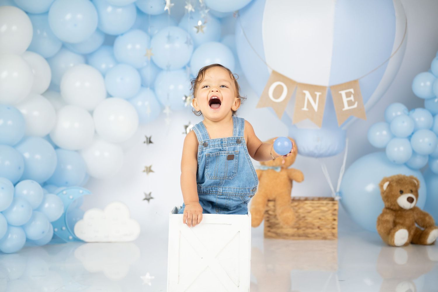 Happy baby in overalls against blue balloon backdrop with teddy bears and ONE banner.