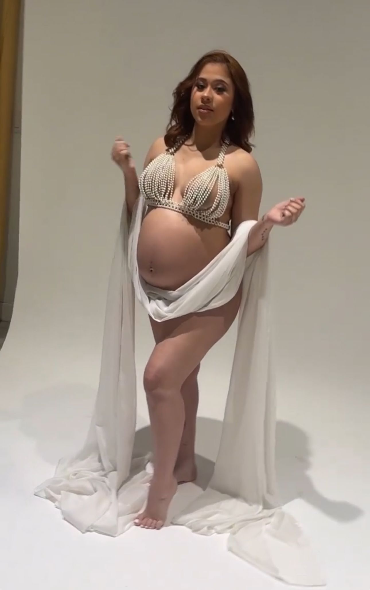 Pregnant woman posing in pearl bikini top and flowing white fabric during a maternity photoshoot.