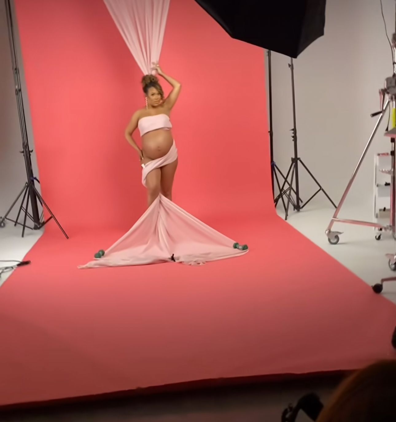 Pregnant woman posing in white draped fabric outfit against pink backdrop in professional photo studio setting.
