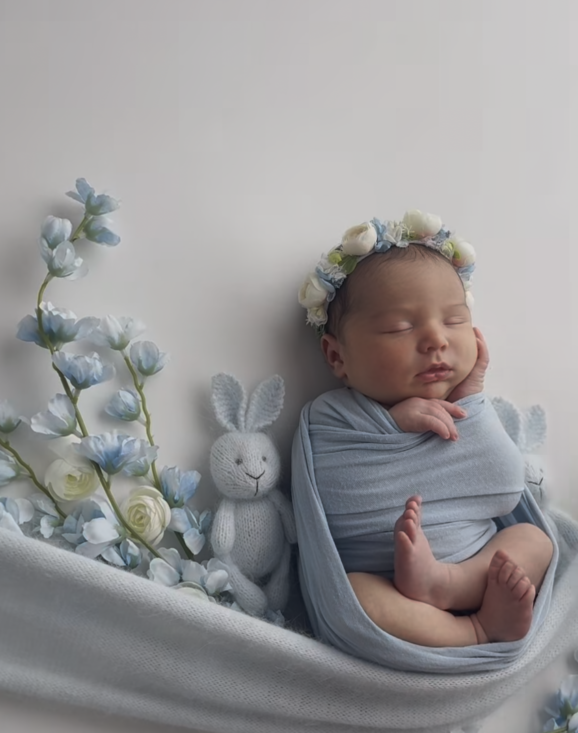 Sleeping newborn baby wrapped in blue swaddle with floral crown, bunny toy, and blue flowers on white background.