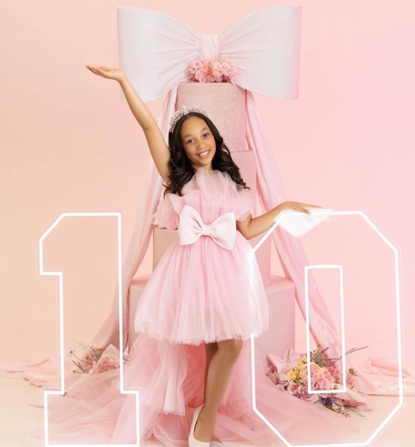 A young girl in a pink tulle dress with a bow poses in front of a large number 10 and giant pink gift bow backdrop.