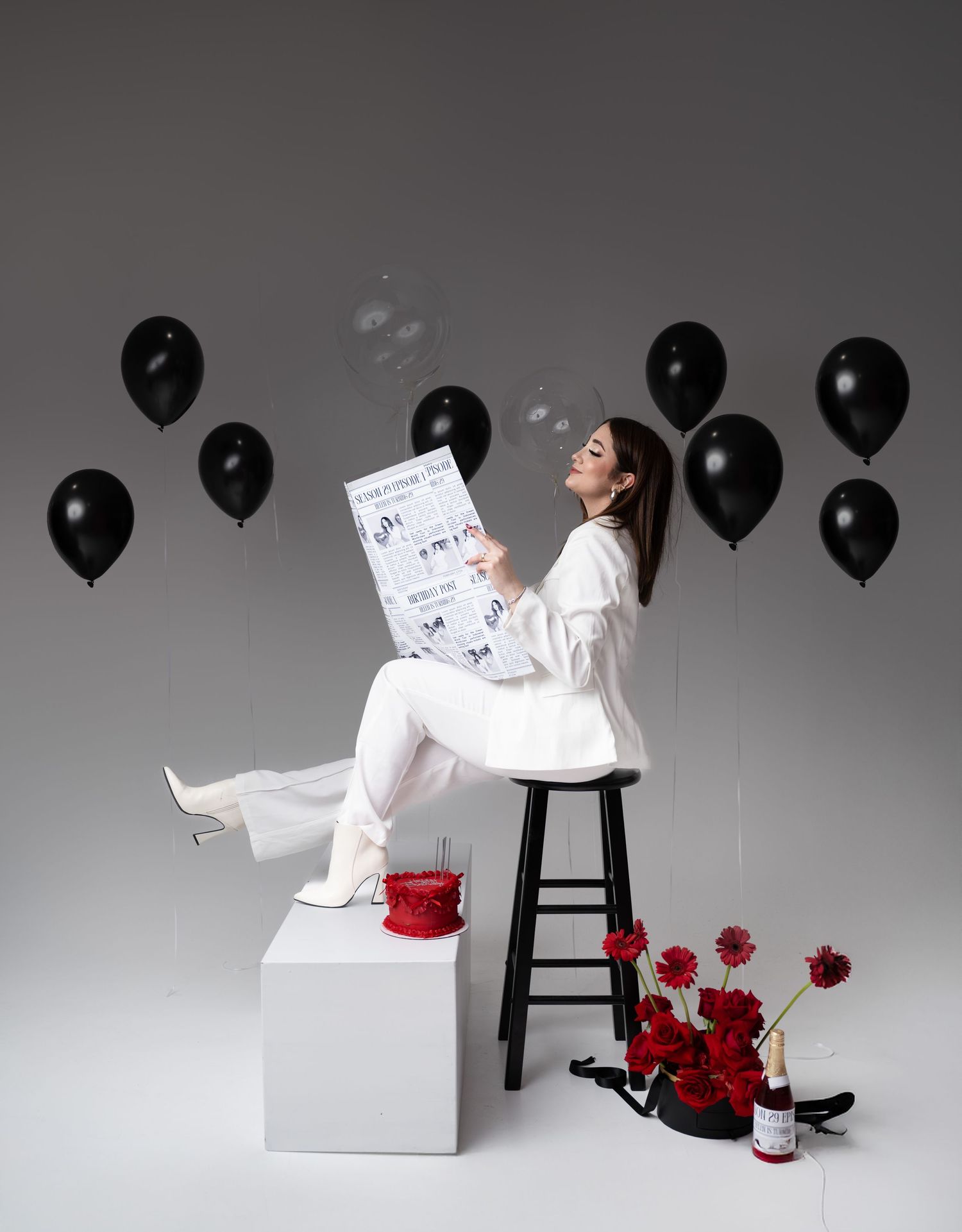 Woman in white outfit sits on stool reading newspaper, surrounded by black balloons and red flowers with champagne bottle.