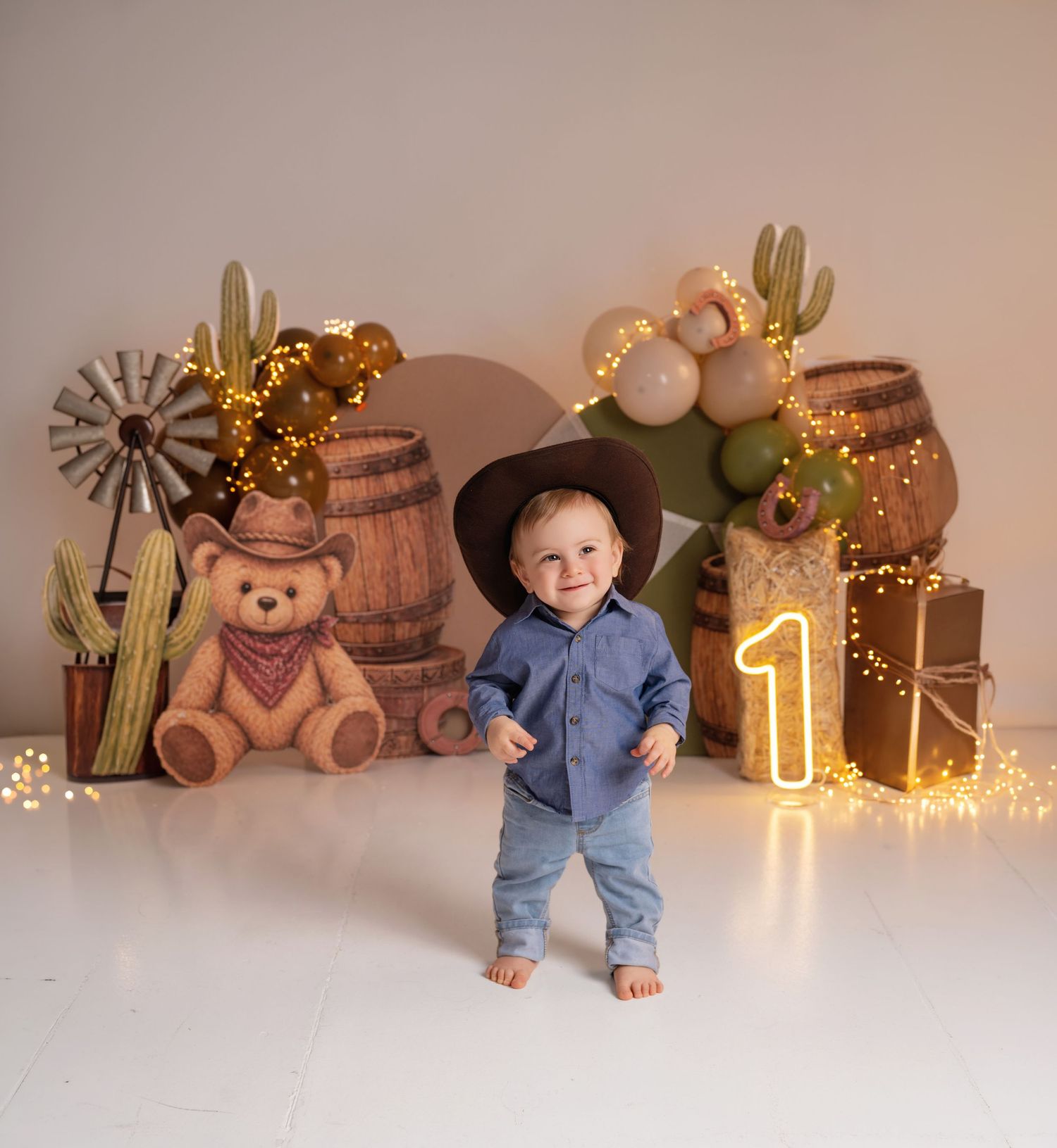 Toddler boy in cowboy hat at Western-themed first birthday party with cactus, barrels, and glowing number one.