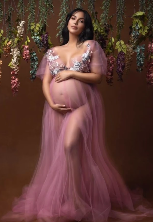 A flowing mauve tulle gown against a floral backdrop creates an ethereal maternity portrait.