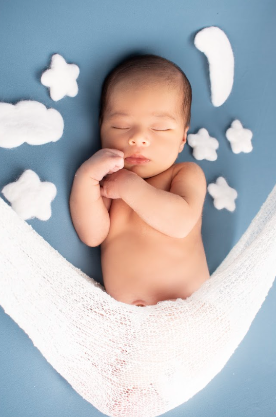 Sleeping newborn baby lying on white lace blanket with paper cloud decorations against blue background.
