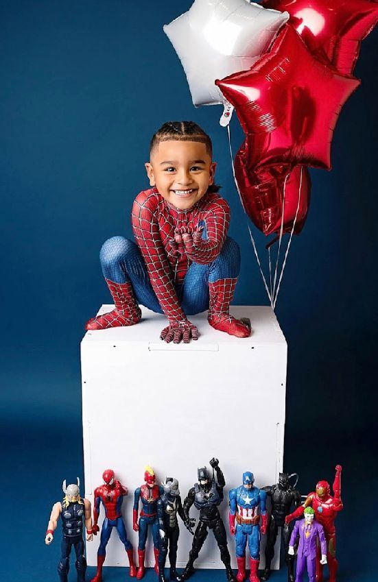 A child in Spider-Man costume poses on a white cube with red star balloons and action figures displayed below.