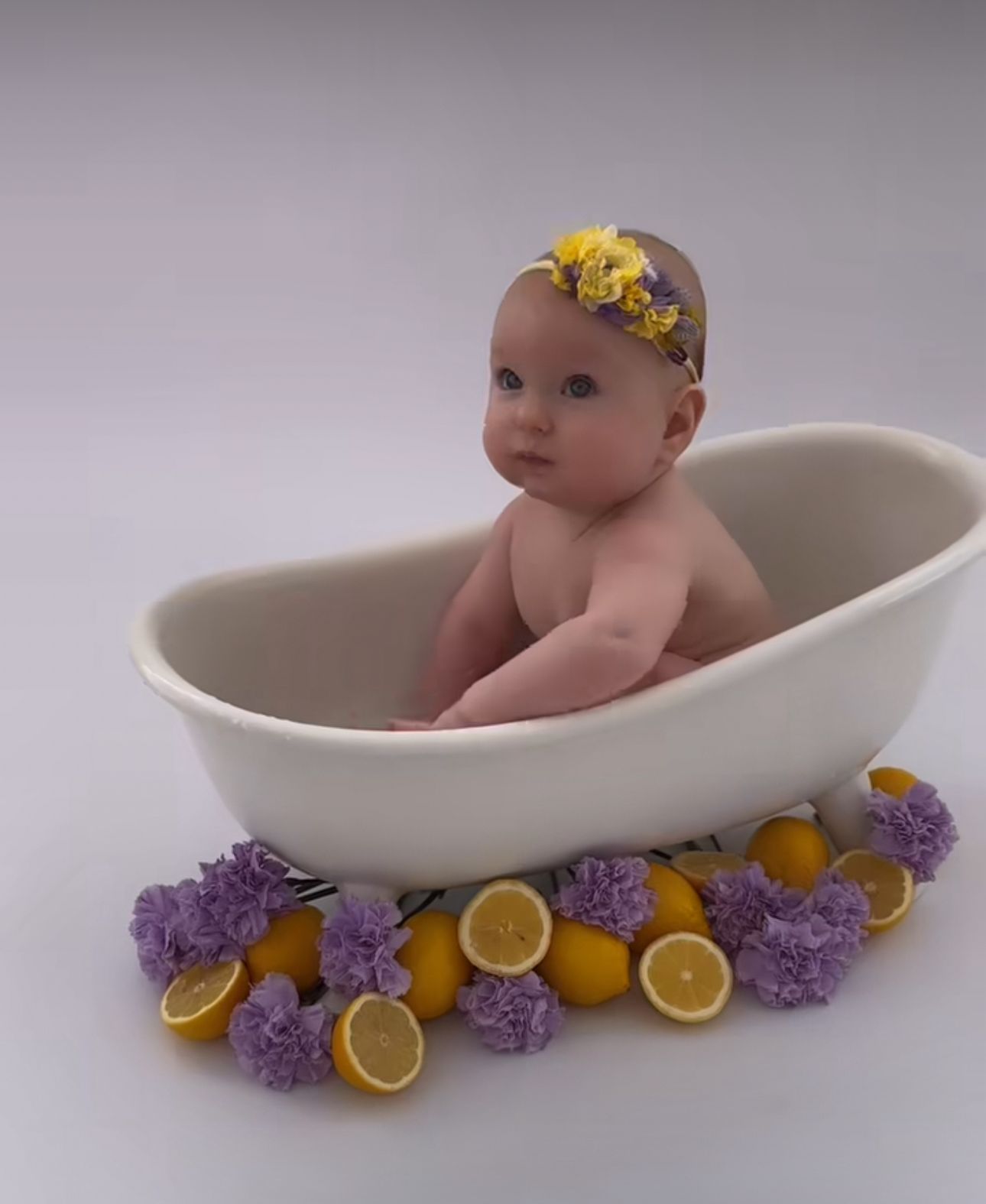 Baby in white bathtub with yellow flower crown, surrounded by purple lilacs and sliced lemons on white background.