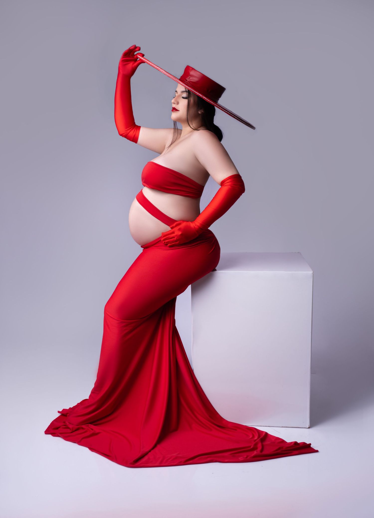 Maternity photo featuring dramatic red gown and hat pose on white block pedestal.