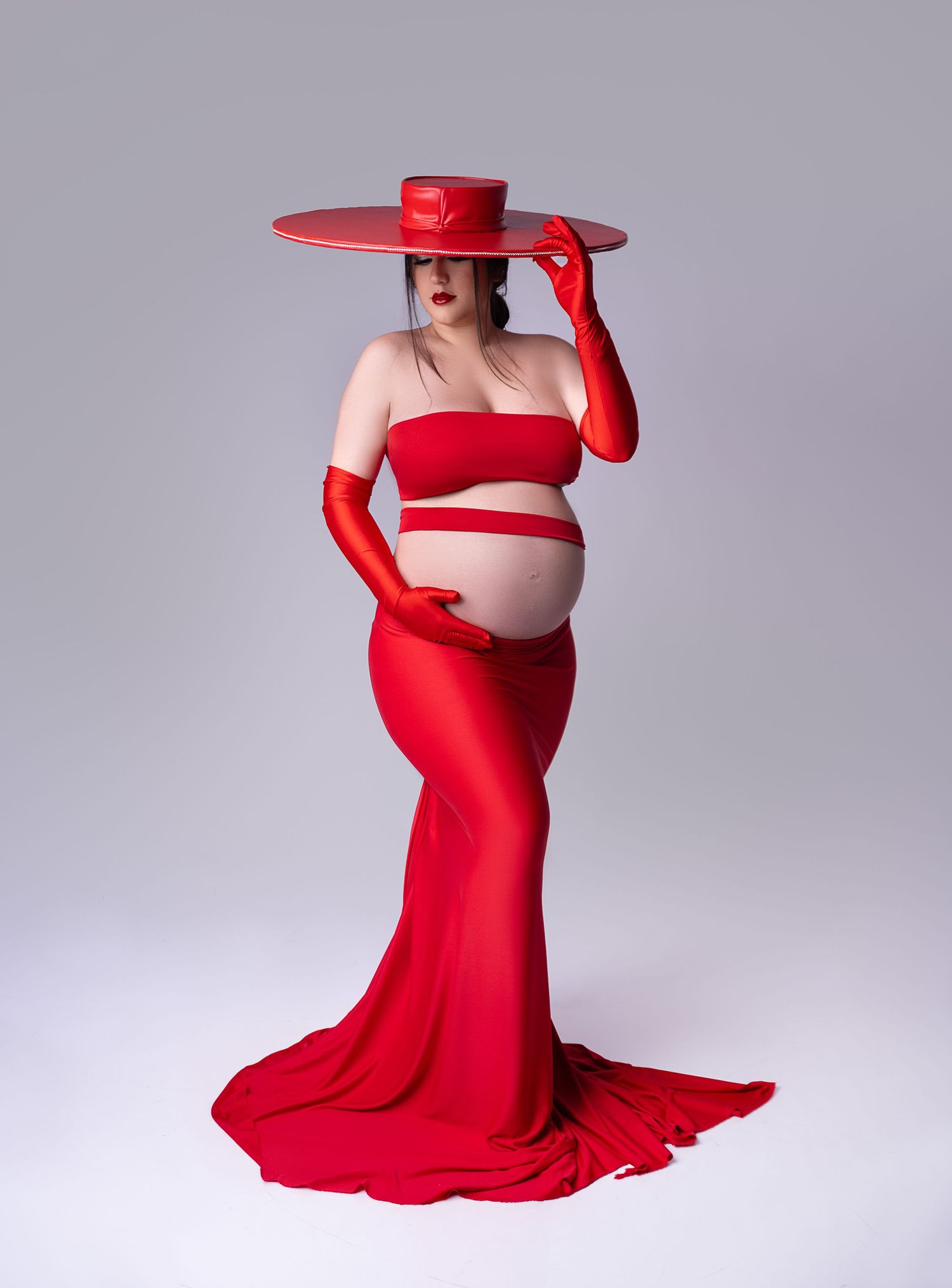Dramatic maternity portrait in flowing red gown with matching wide-brimmed hat against gray background.