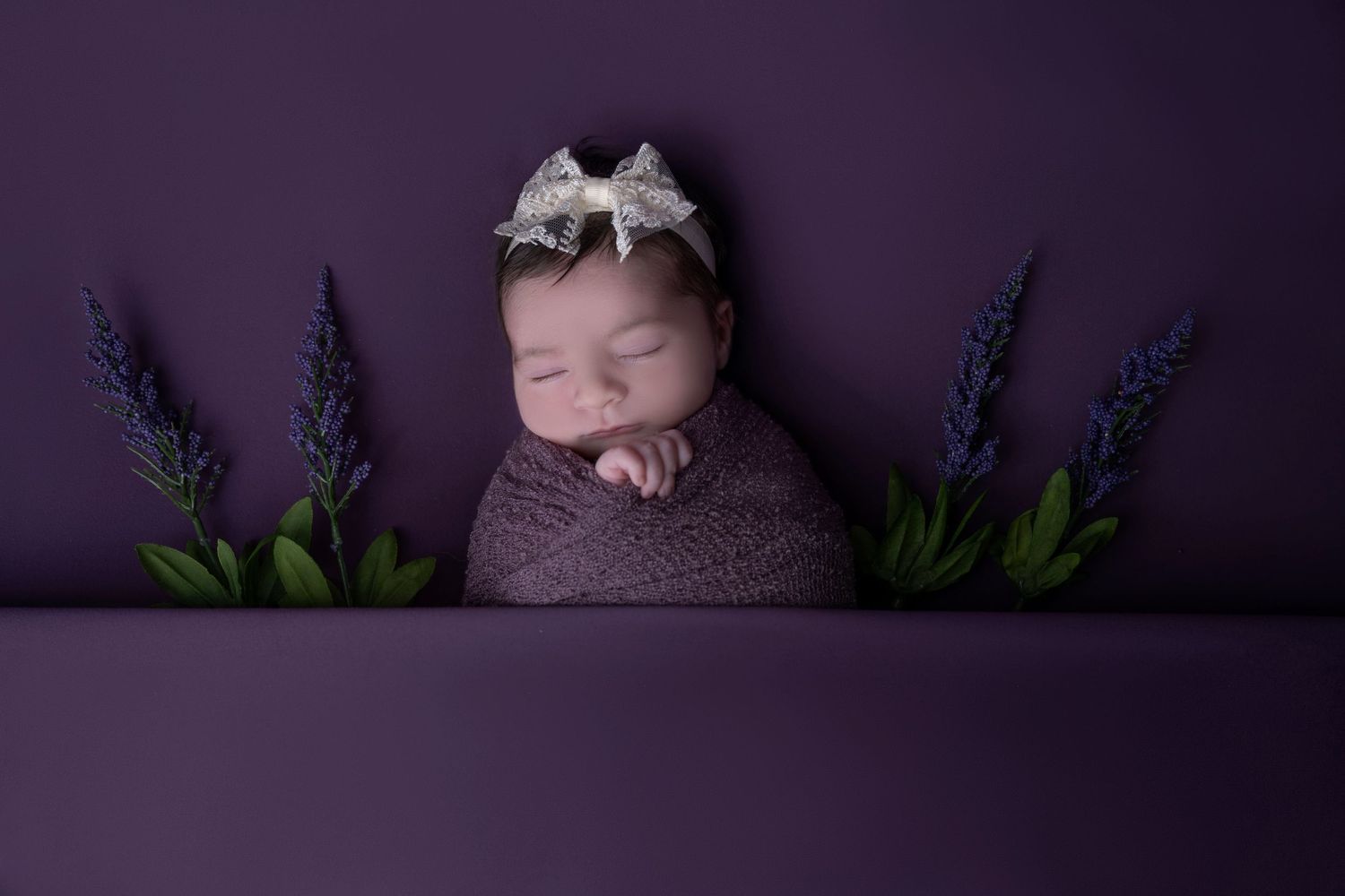 Sleeping newborn wrapped in purple swaddle with lavender flowers and bow headband.